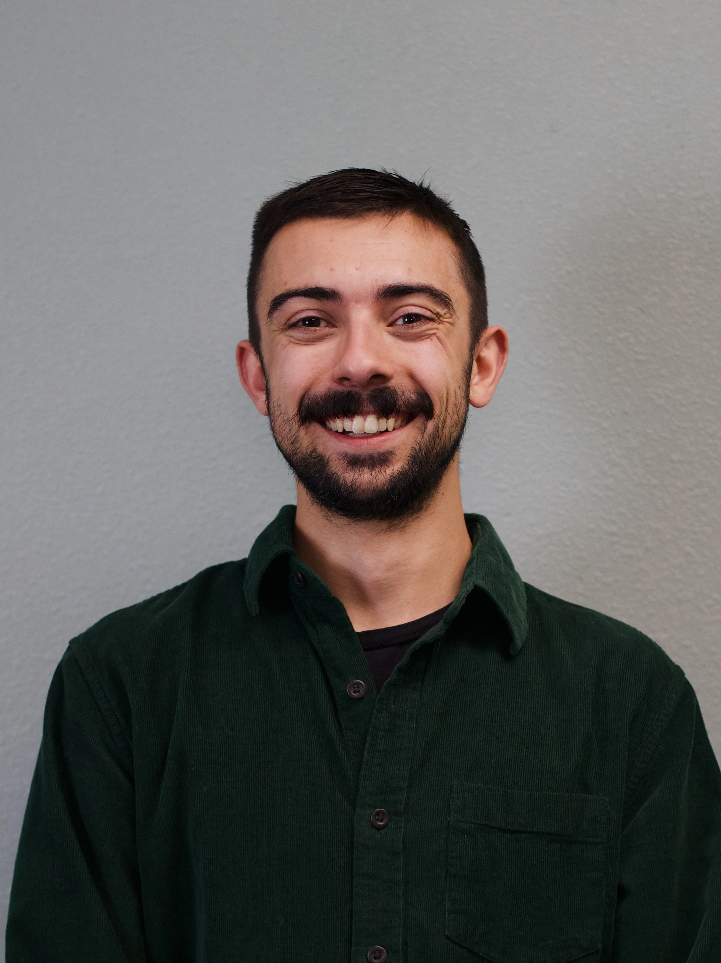 A young man with dark hair and a beard, smiling, standing in front of a red brick wall, wearing a dark, polka-dotted shirt.