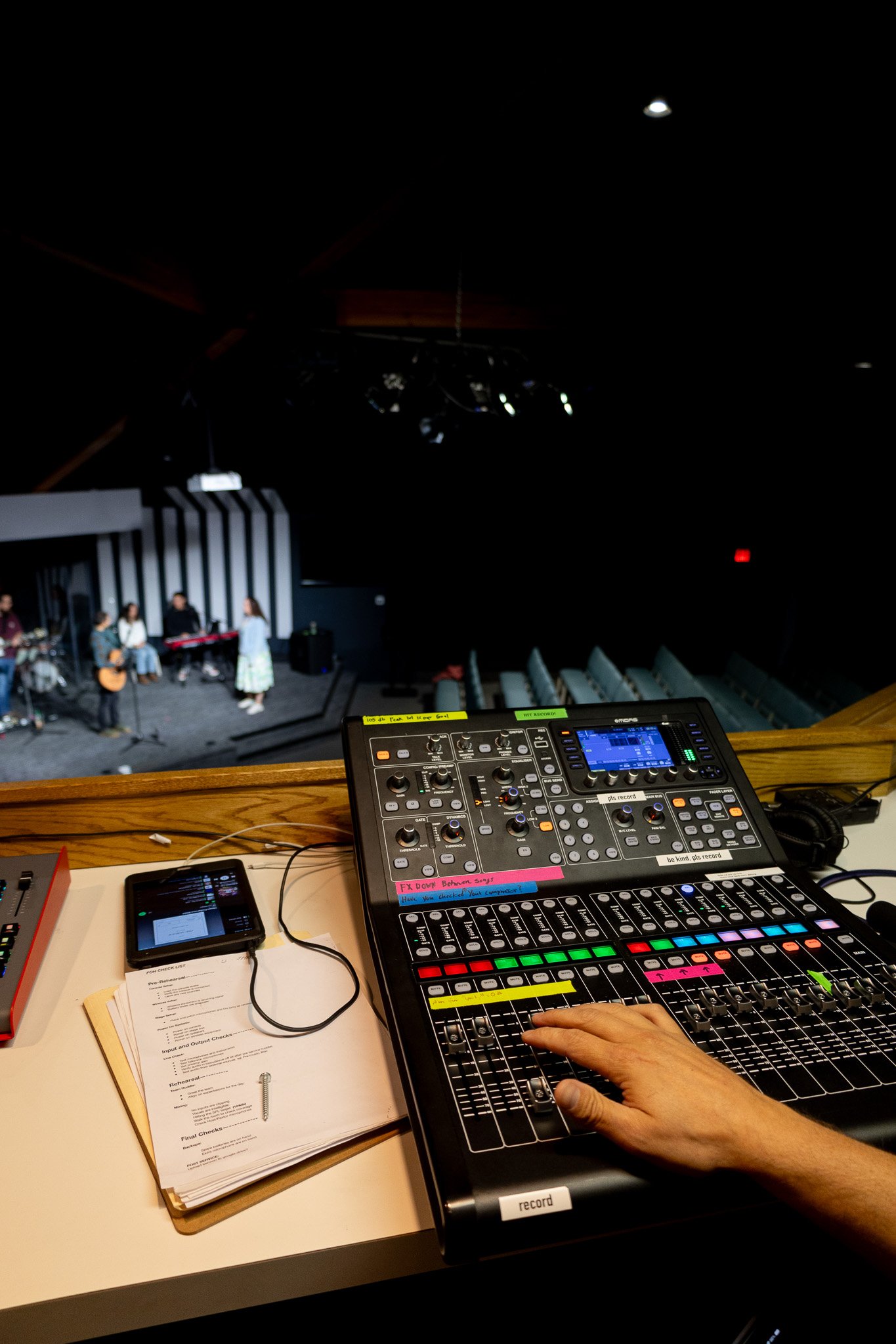View of a live music rehearsal or performance from the sound booth, showing a sound mixer with a person's hand adjusting controls, a pull list, and a blurry stage with musicians and singers in the background.