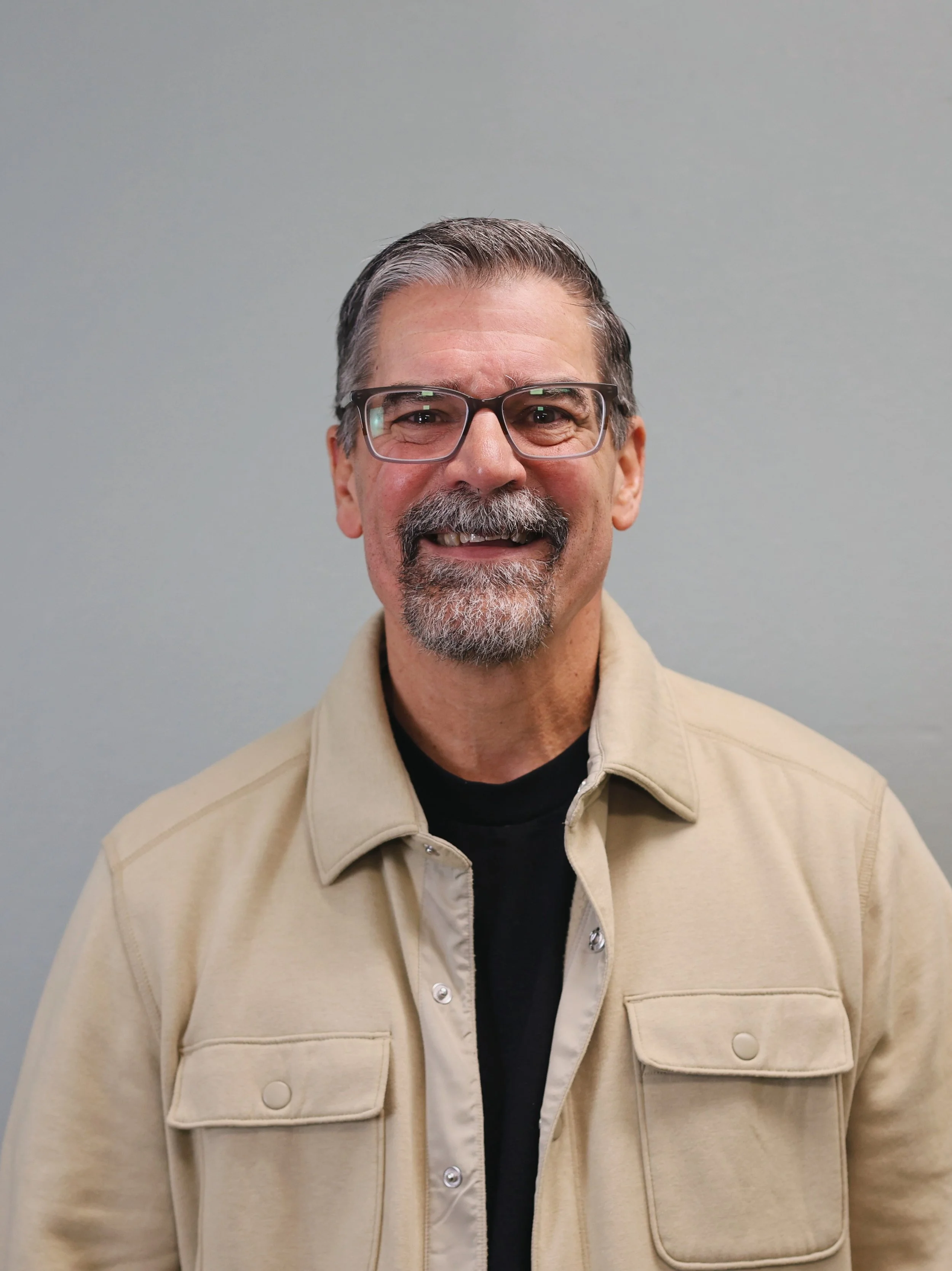 A smiling middle-aged man with gray hair and a beard, wearing a gray shirt, standing in front of a red brick wall.