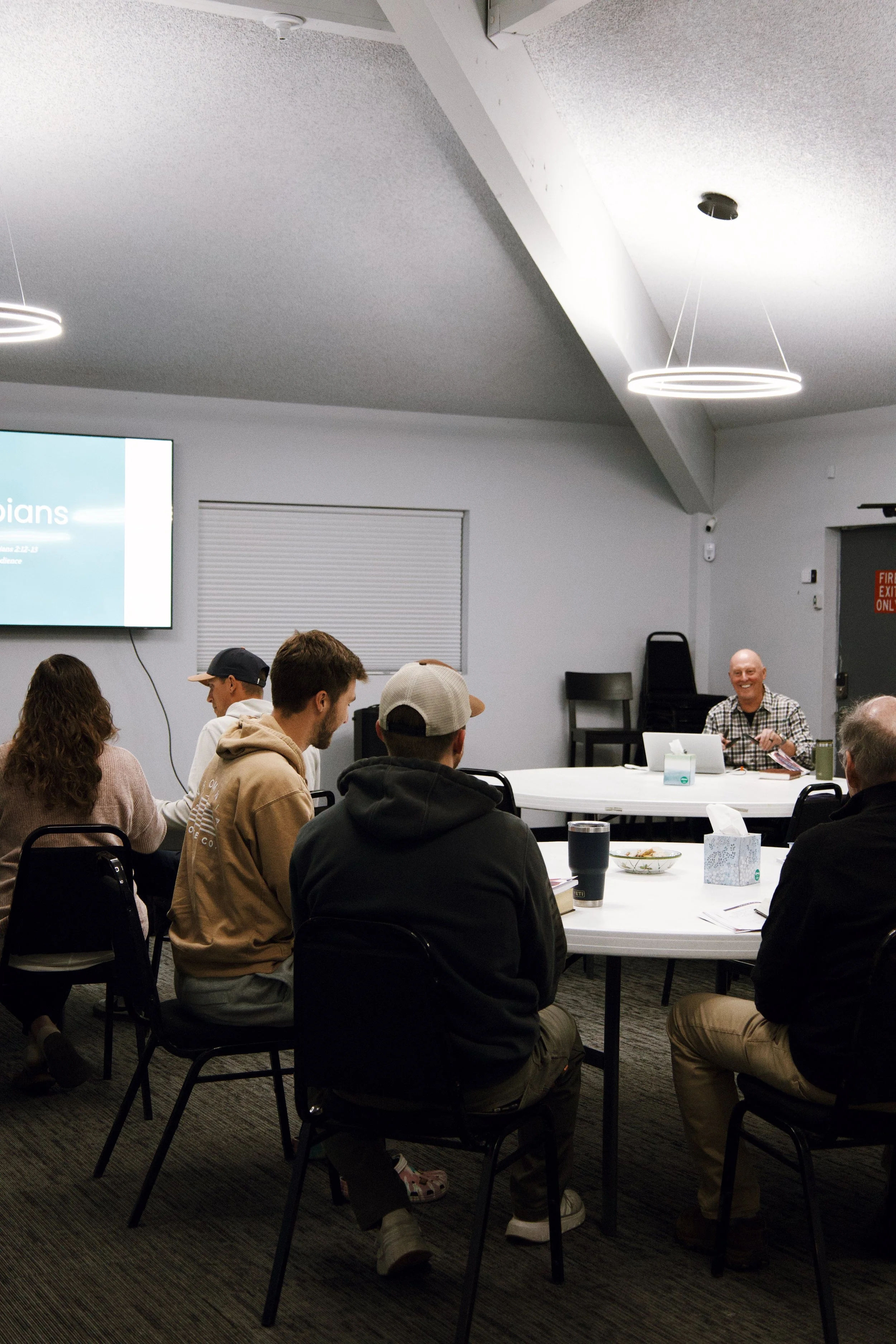 Group of people attending a presentation or meeting in a conference room, with a presenter at the front and a large screen displaying a slide.