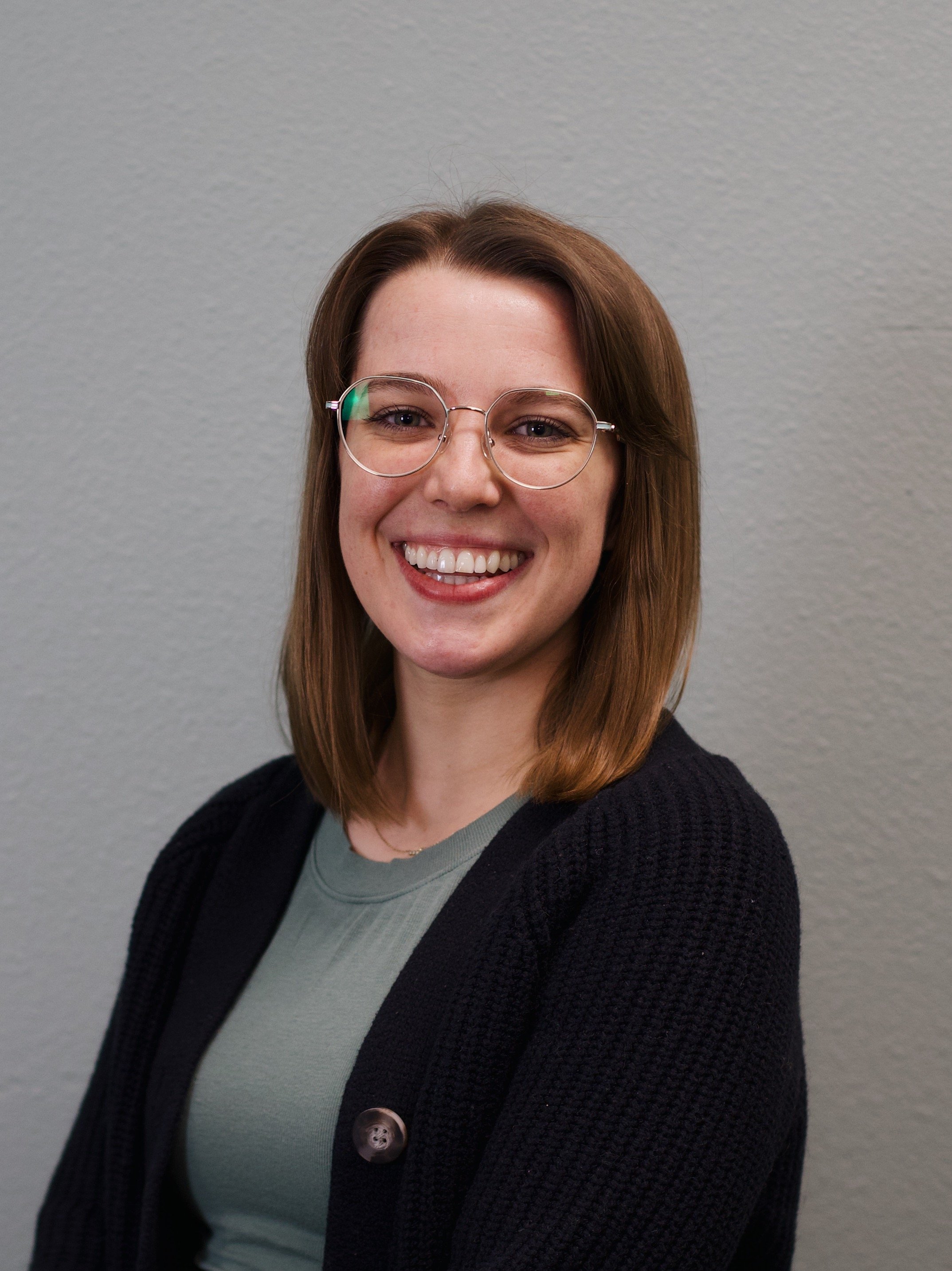 A young woman with shoulder-length brown hair, glasses, and a big smile standing in front of a red brick wall.