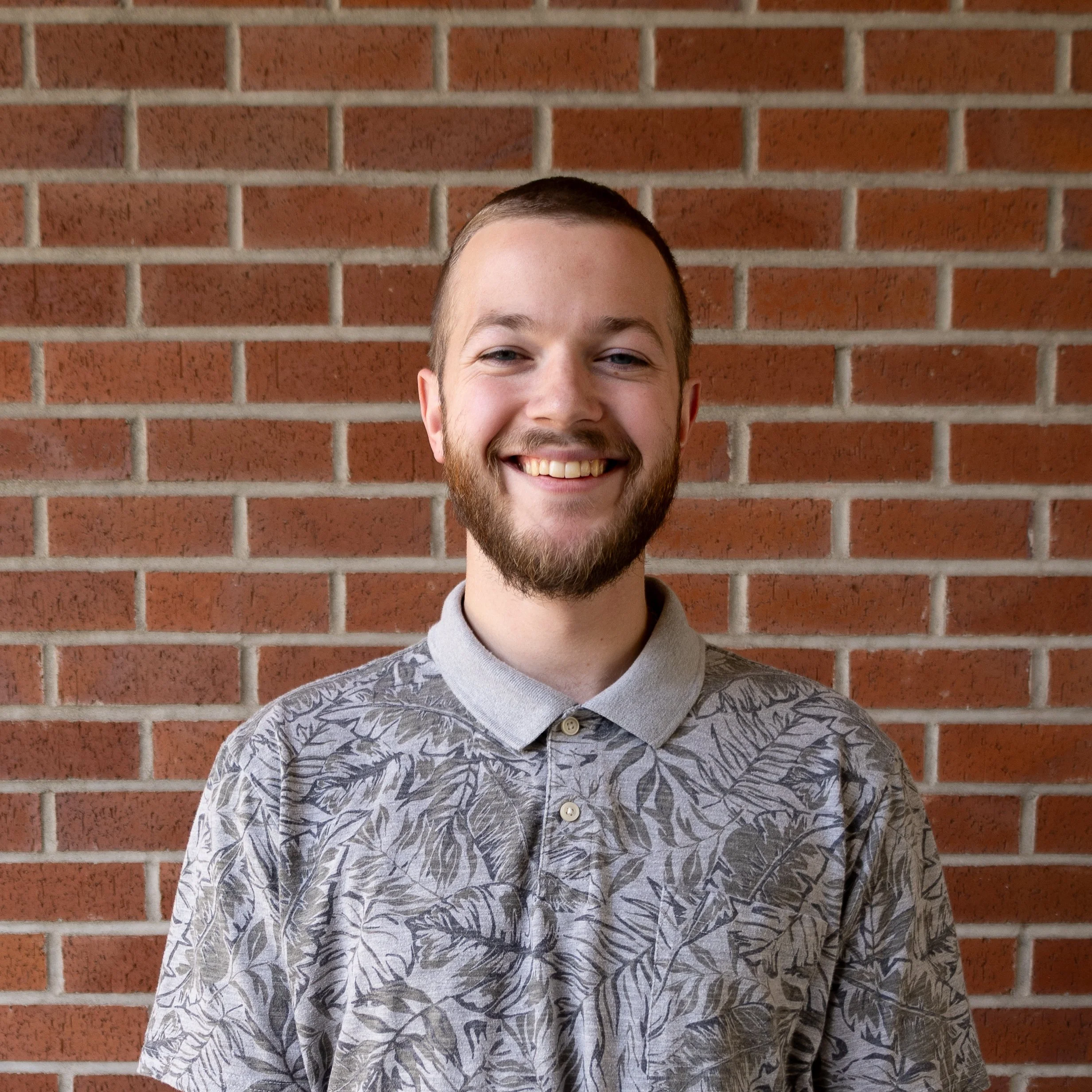 A young man with a beard smiling in front of a brick wall, wearing a patterned grey shirt with a collar.