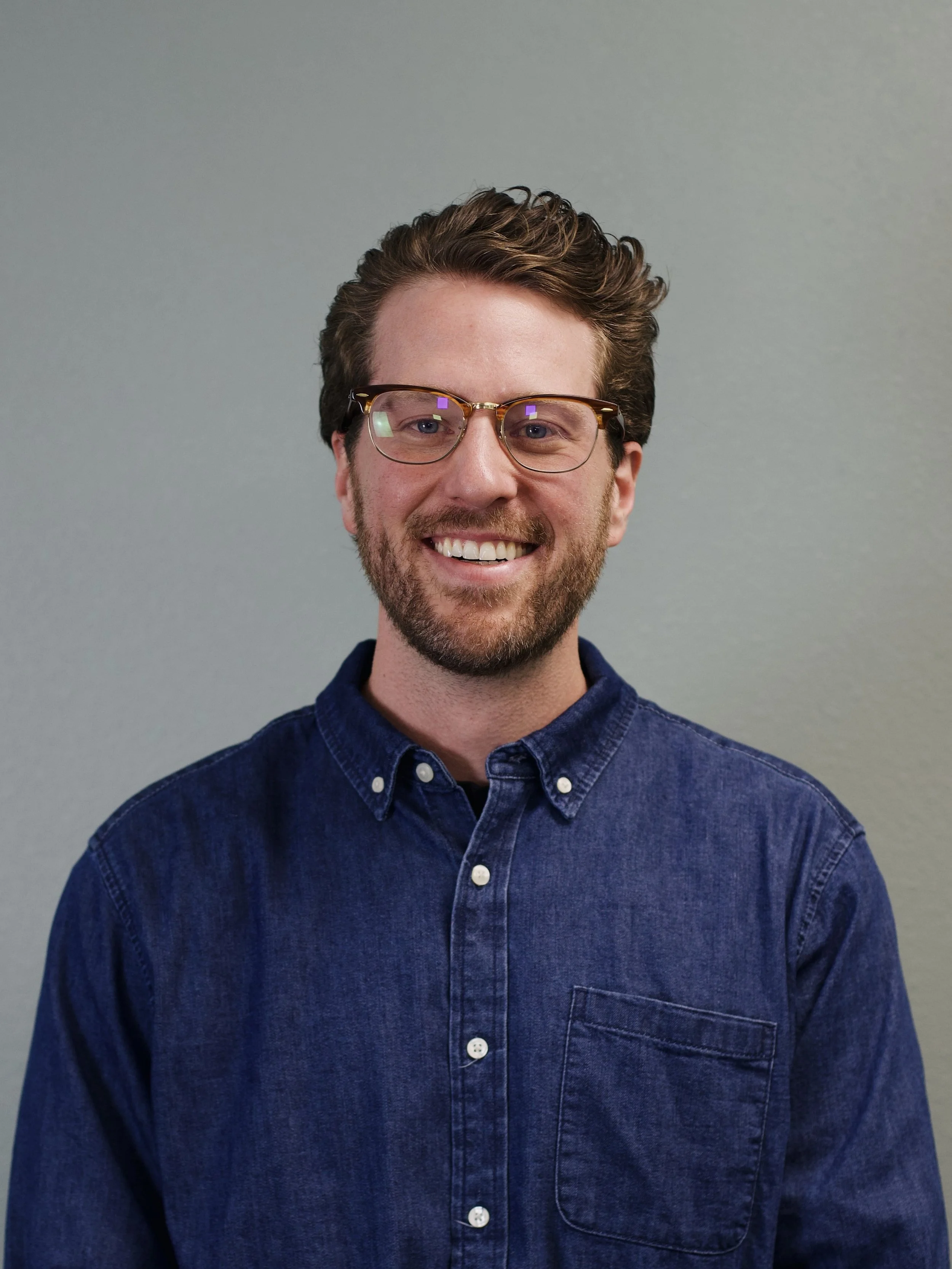 A young man with wavy brown hair, glasses, and a beard, smiling, standing in front of a brick wall, wearing a short-sleeved, striped button-up shirt.