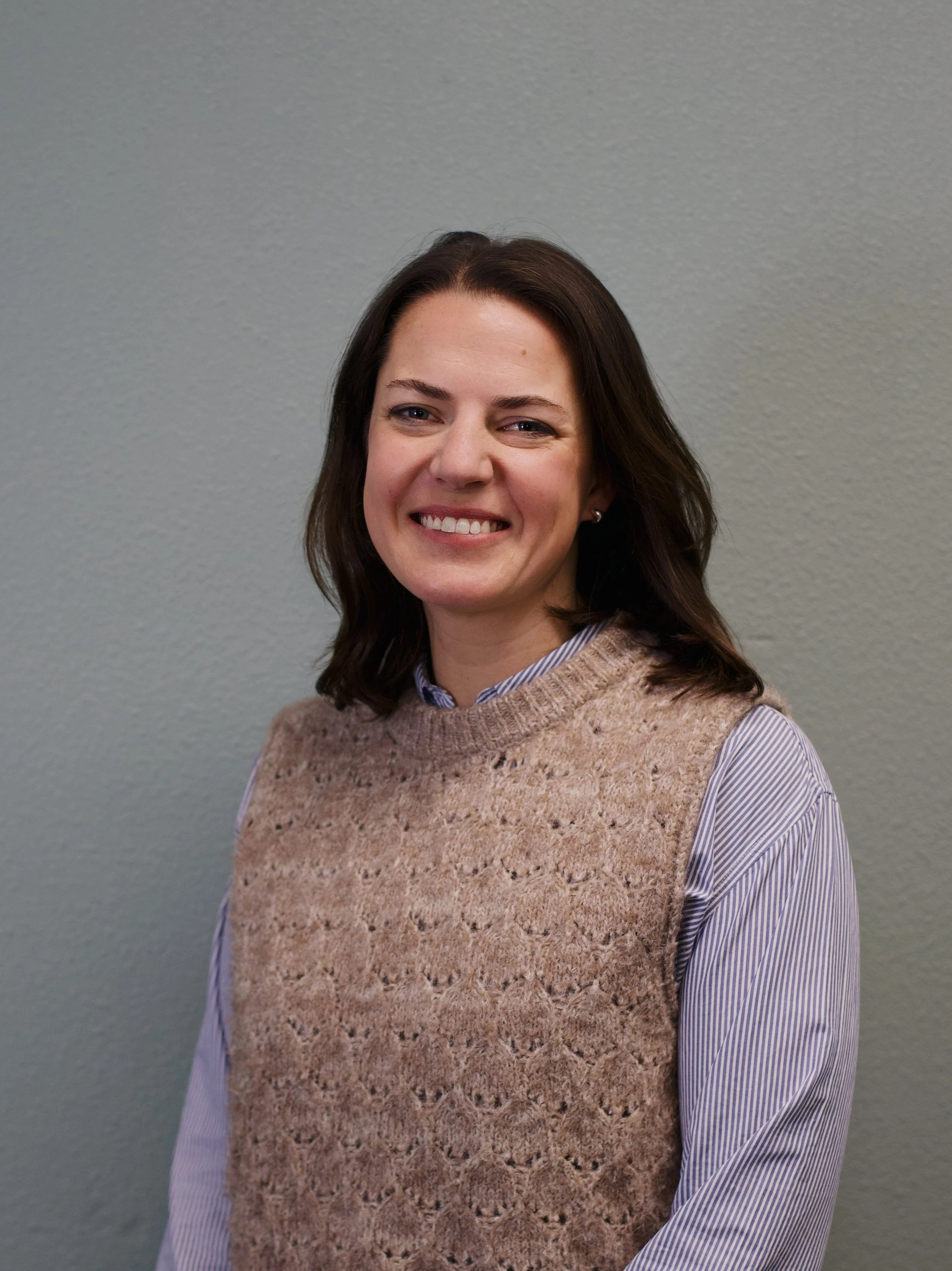 A woman with shoulder-length dark brown hair, smiling, wearing a brown t-shirt, standing in front of a red brick wall.