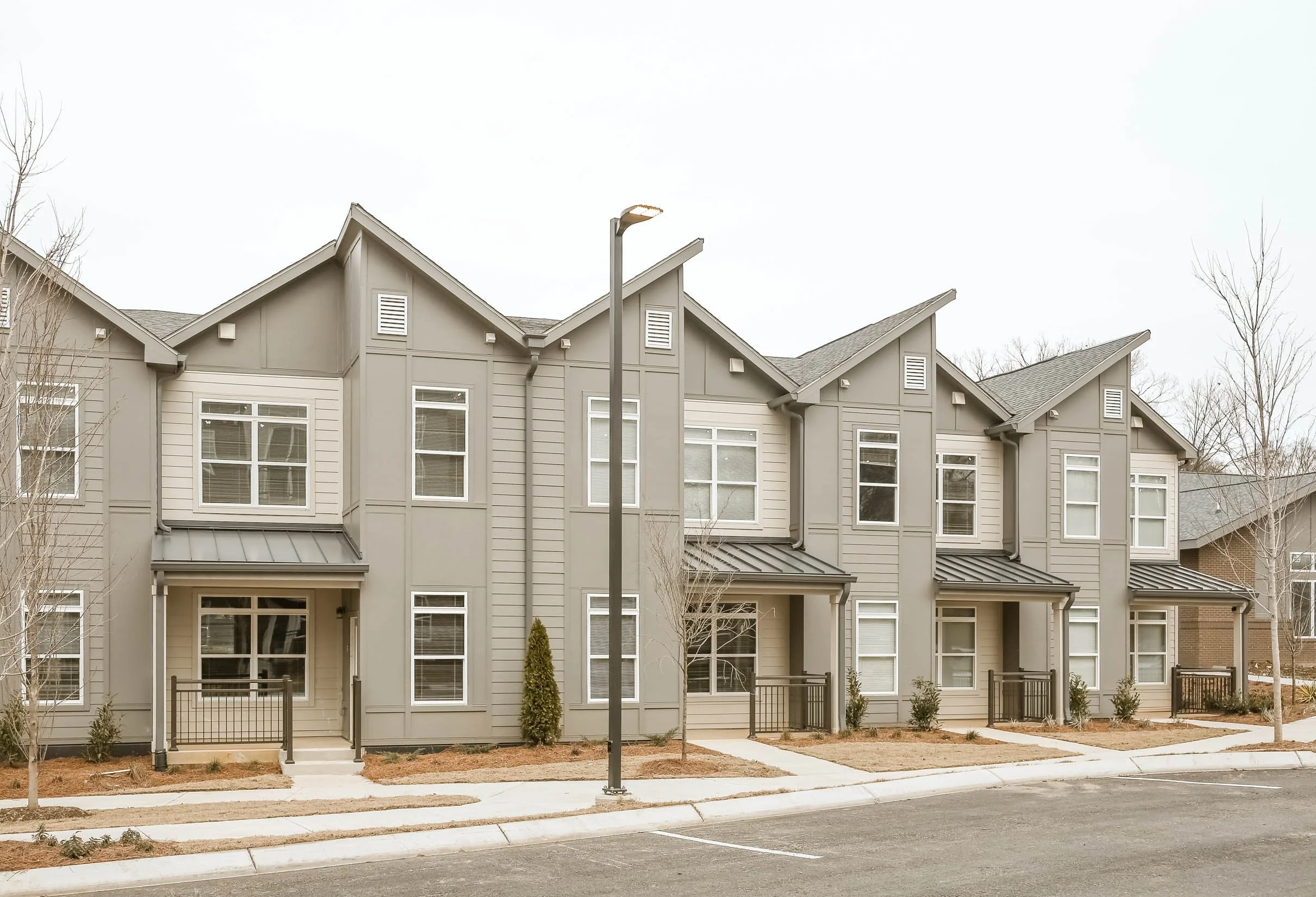 A row of modern townhouse apartments with gray siding, multiple windows, small front porches, and landscaped areas, under an overcast sky.