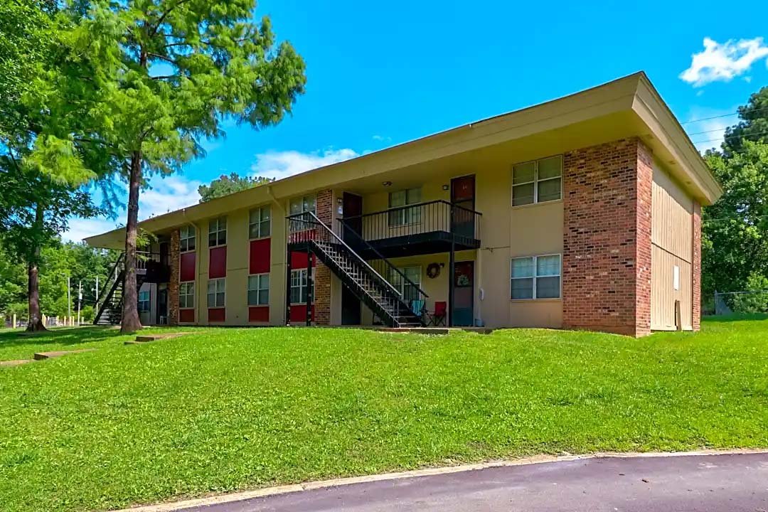 A two-story apartment building with a brick and beige siding exterior, black outdoor stairs, green grass, and trees under a blue sky with some clouds.