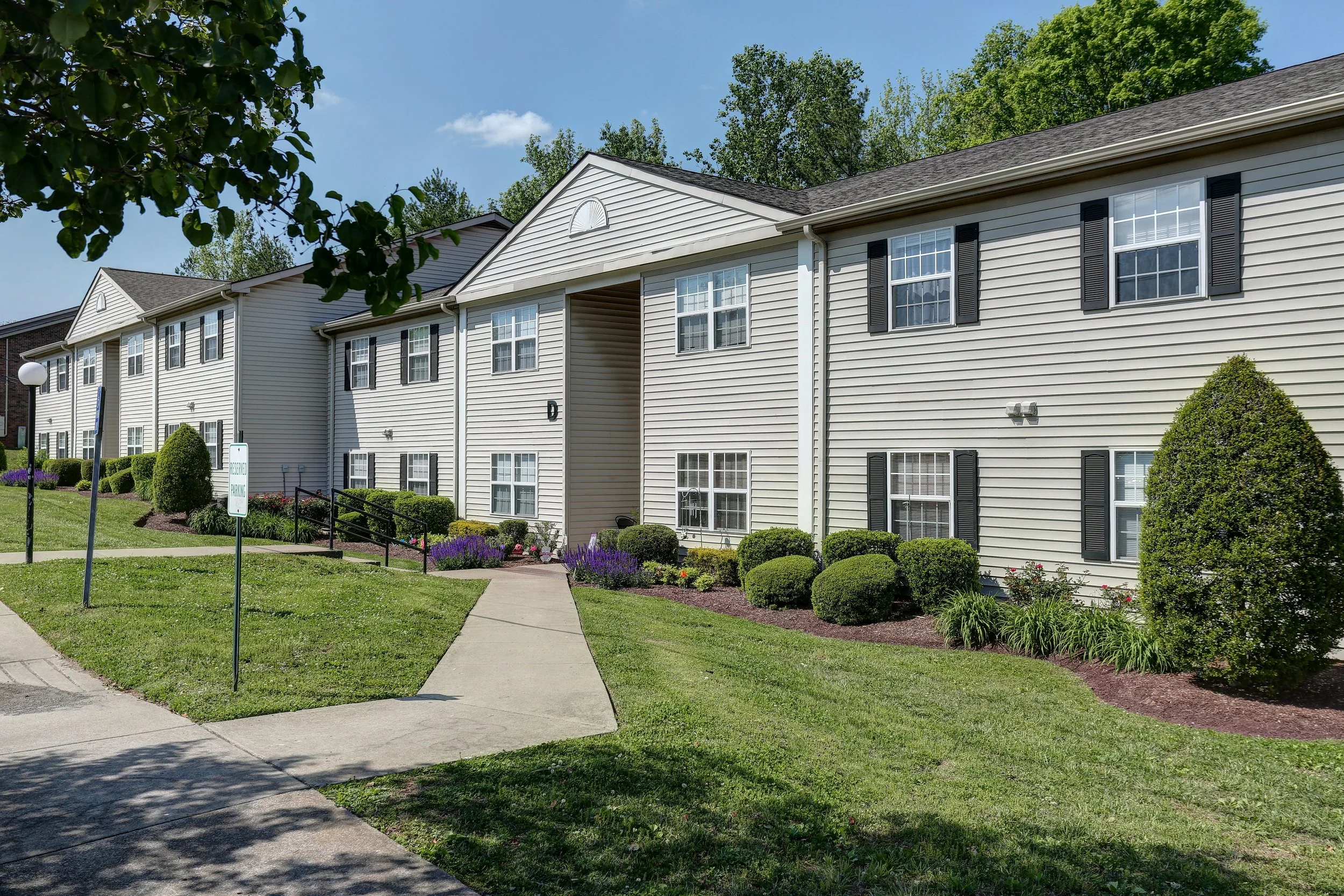 A two-story residential apartment building with beige siding, black shutters, and white window frames, surrounded by well-maintained bushes and green lawn, under a blue sky with some clouds.