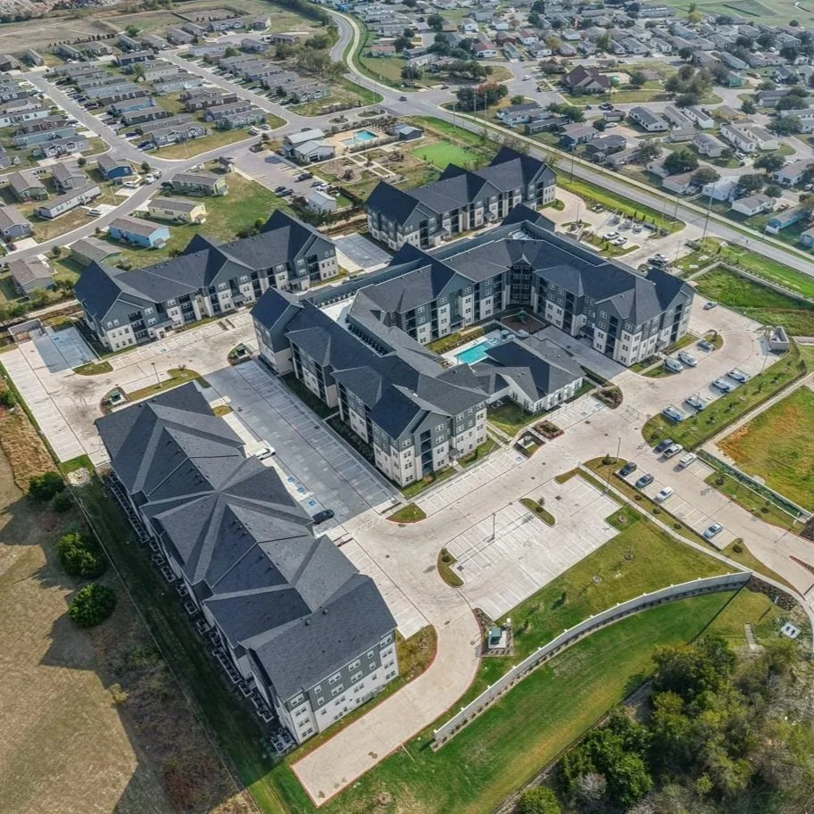 Multi-story apartment complex with parked cars and people walking outside on a sunny day.