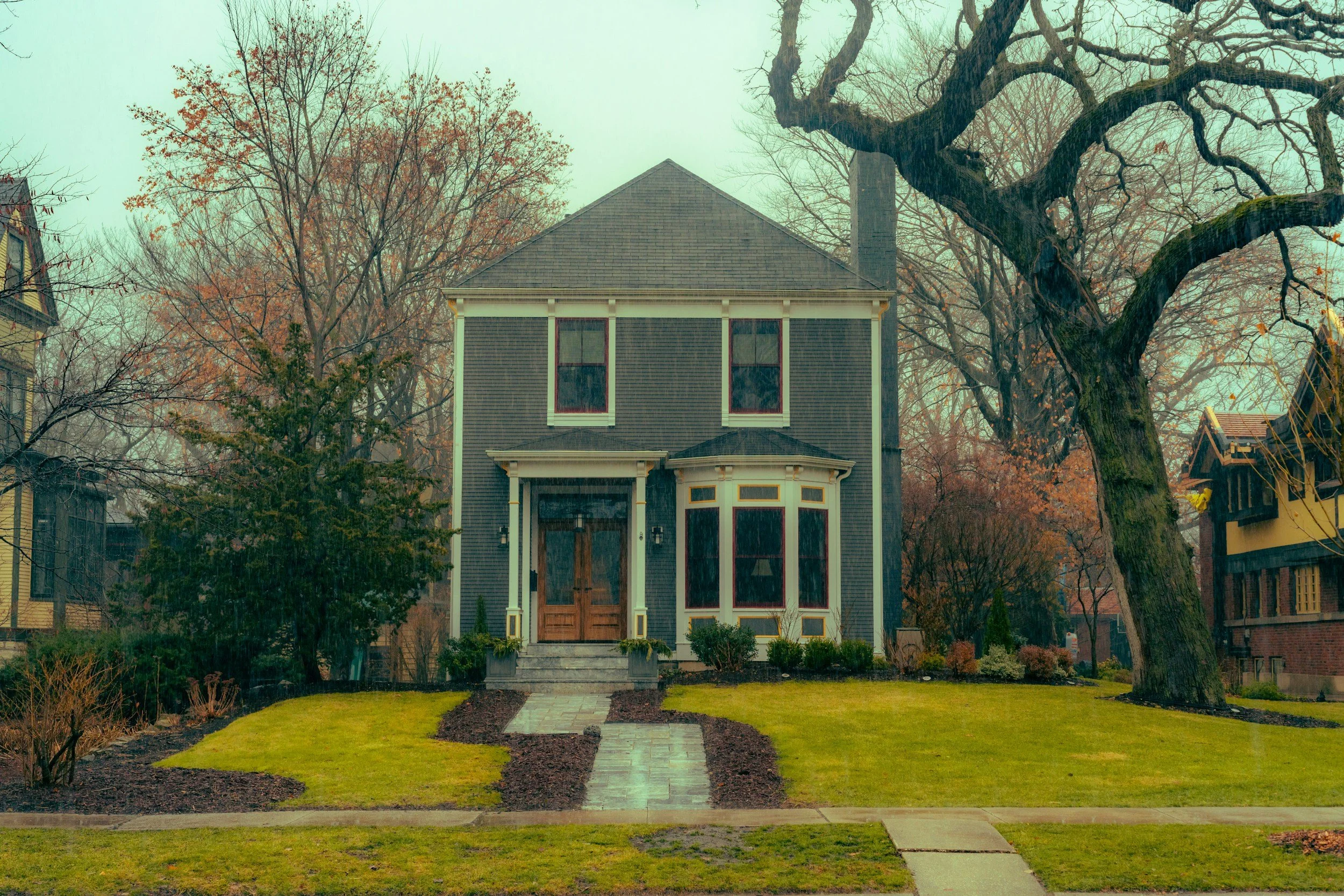 A 2 story house with blue siding, and a brown wood door, with a bay window to the right.