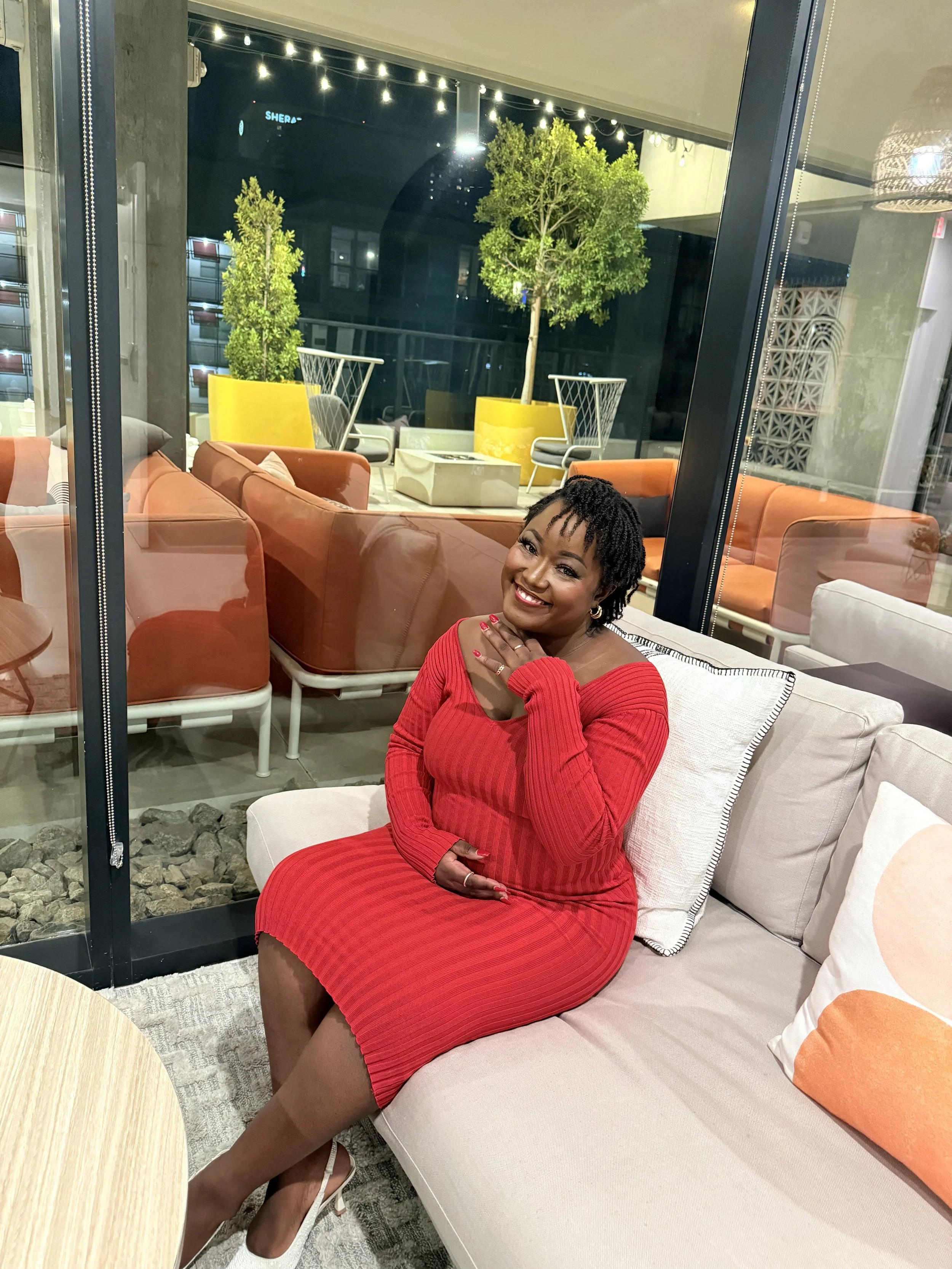 A woman in a red dress sitting on a beige sofa inside a modern cafe or lounge, smiling and touching her face, with large windows behind her showing outdoor seating with plants and string lights at night.