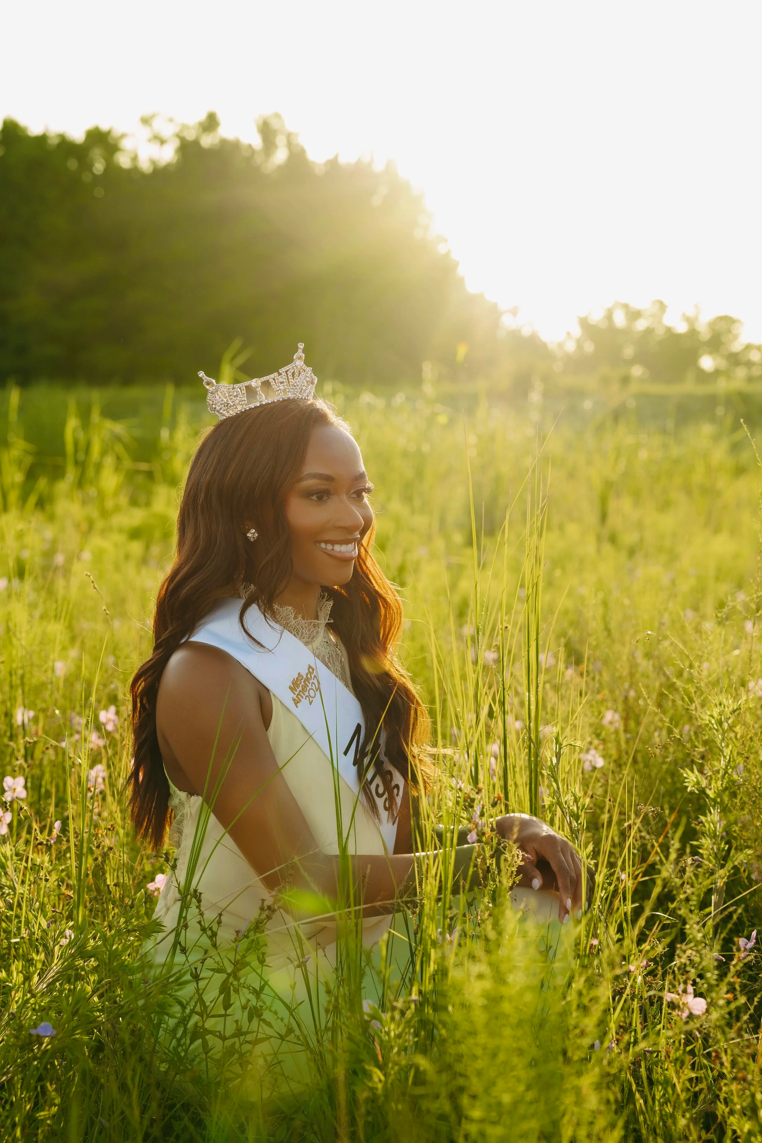 A woman wearing a crown and sash, sitting among wildflowers and tall grass in a field during golden hour sunlight.