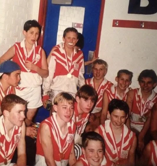 A group of young boys and a girl in red and white striped sports uniforms, some with medals, posing for a photo in a gym or sports facility.