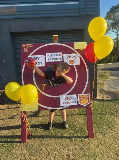 Child standing behind a large target with balloons on the side, holding a football, outdoors near a building and trees.