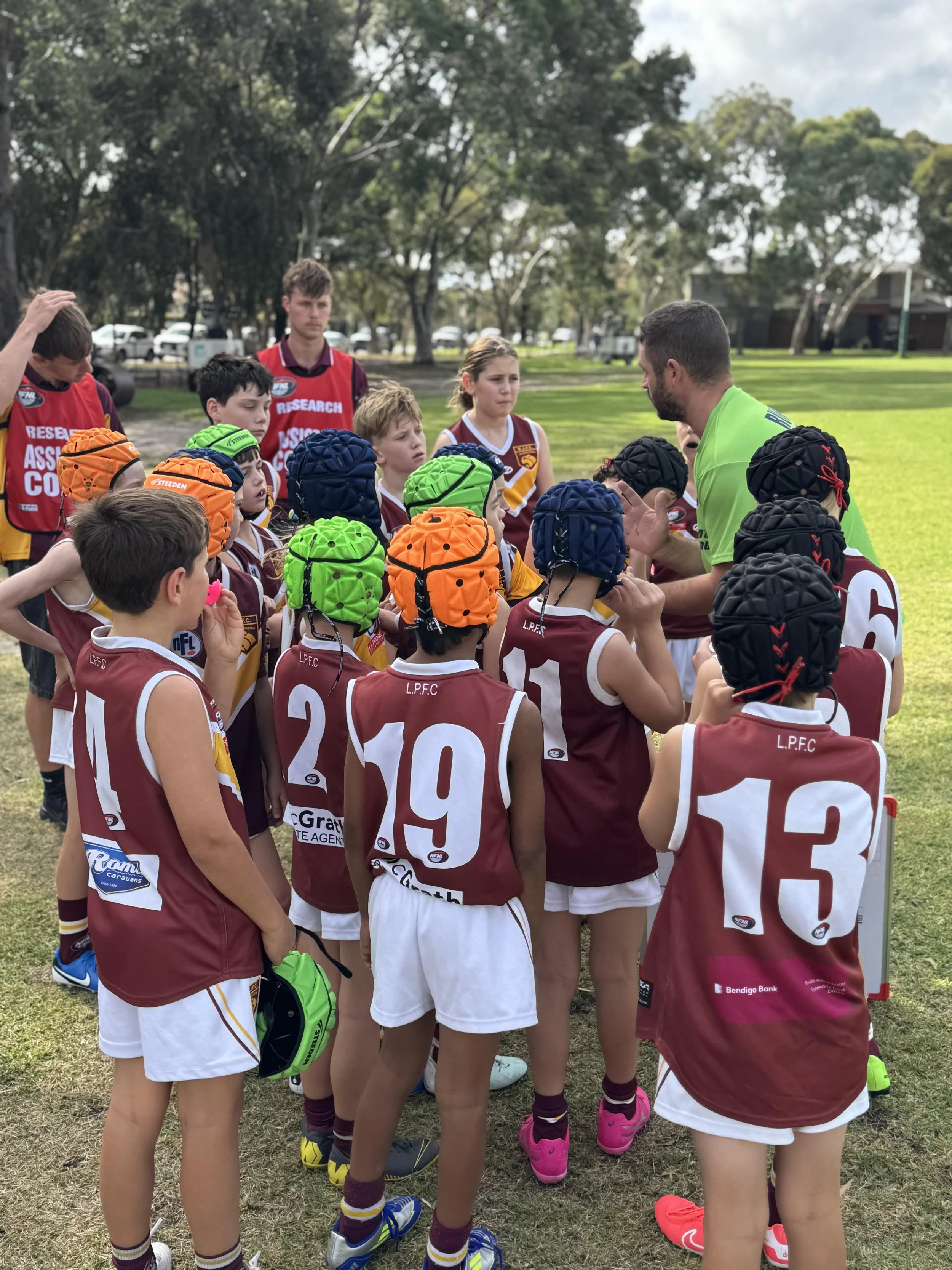 Young Australian rules football players in maroon and white uniforms listening to their coach during a game.