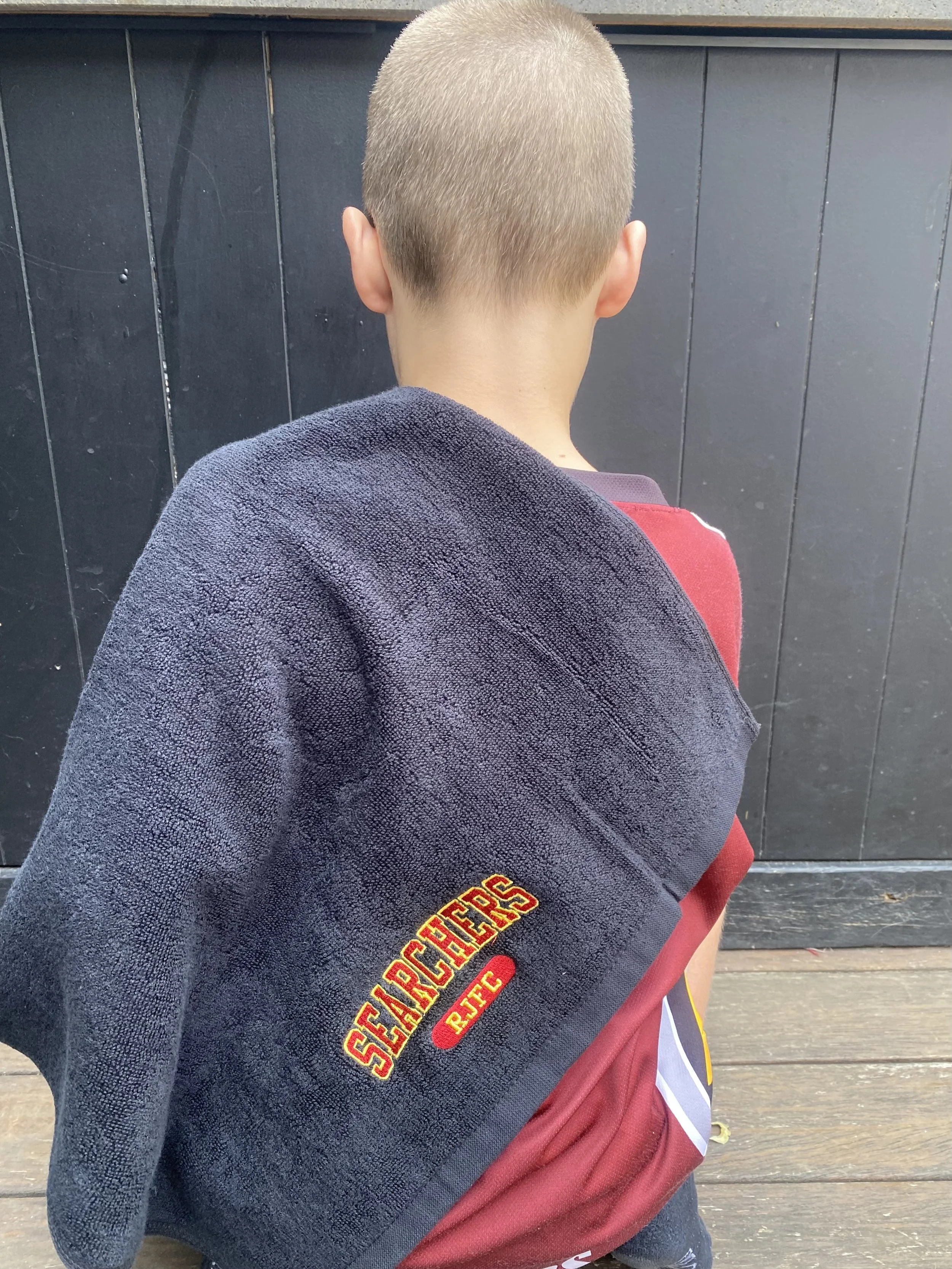 Back of a young child with a shaved head, wearing a dark towel with the word Searchers embroidered on it, standing in front of a black wooden fence.