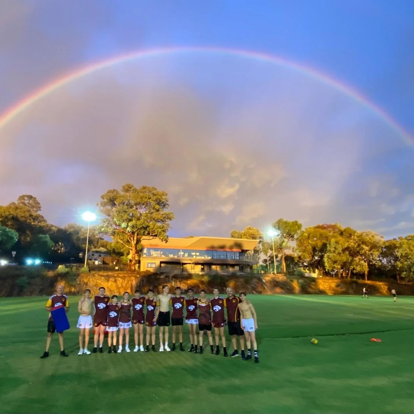 A group of junior football players standing at Research Park at dusk, with a rainbow and clouds in the sky, and a building with trees in the background.