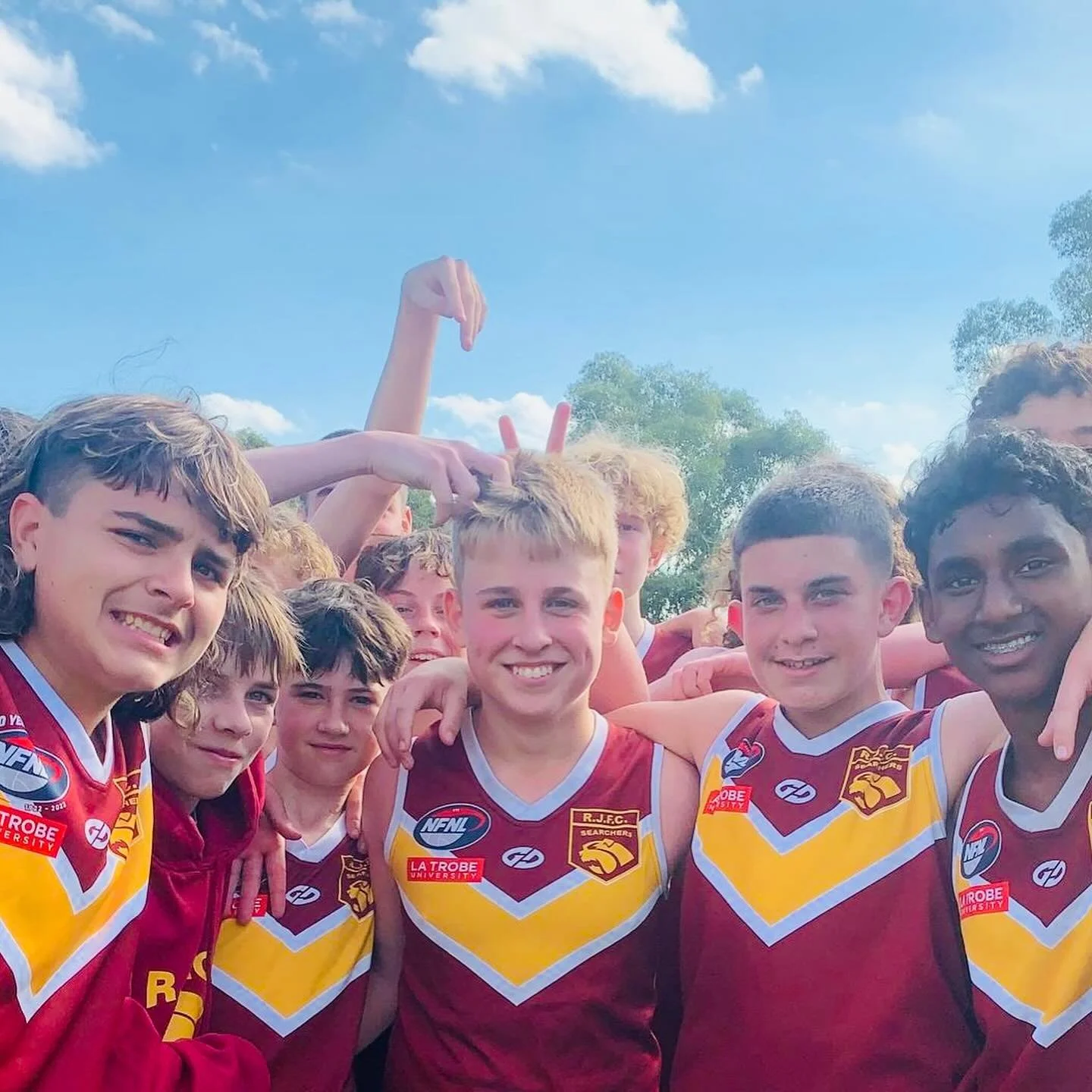 Group of young male athletes in maroon and gold sports uniforms smiling outdoors on a sunny day.