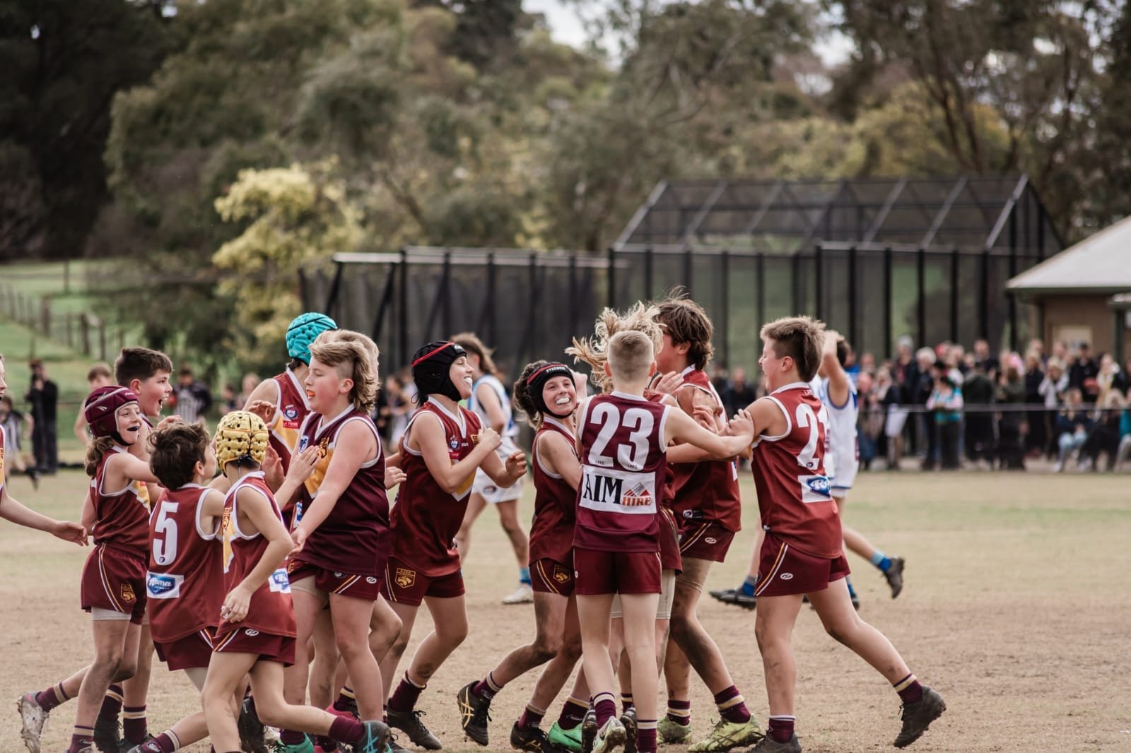 A group of young boys and girls in maroon and yellow sports uniforms celebrating on a grassy field, with some wearing helmets, in a park with trees and a crowd of spectators in the background.