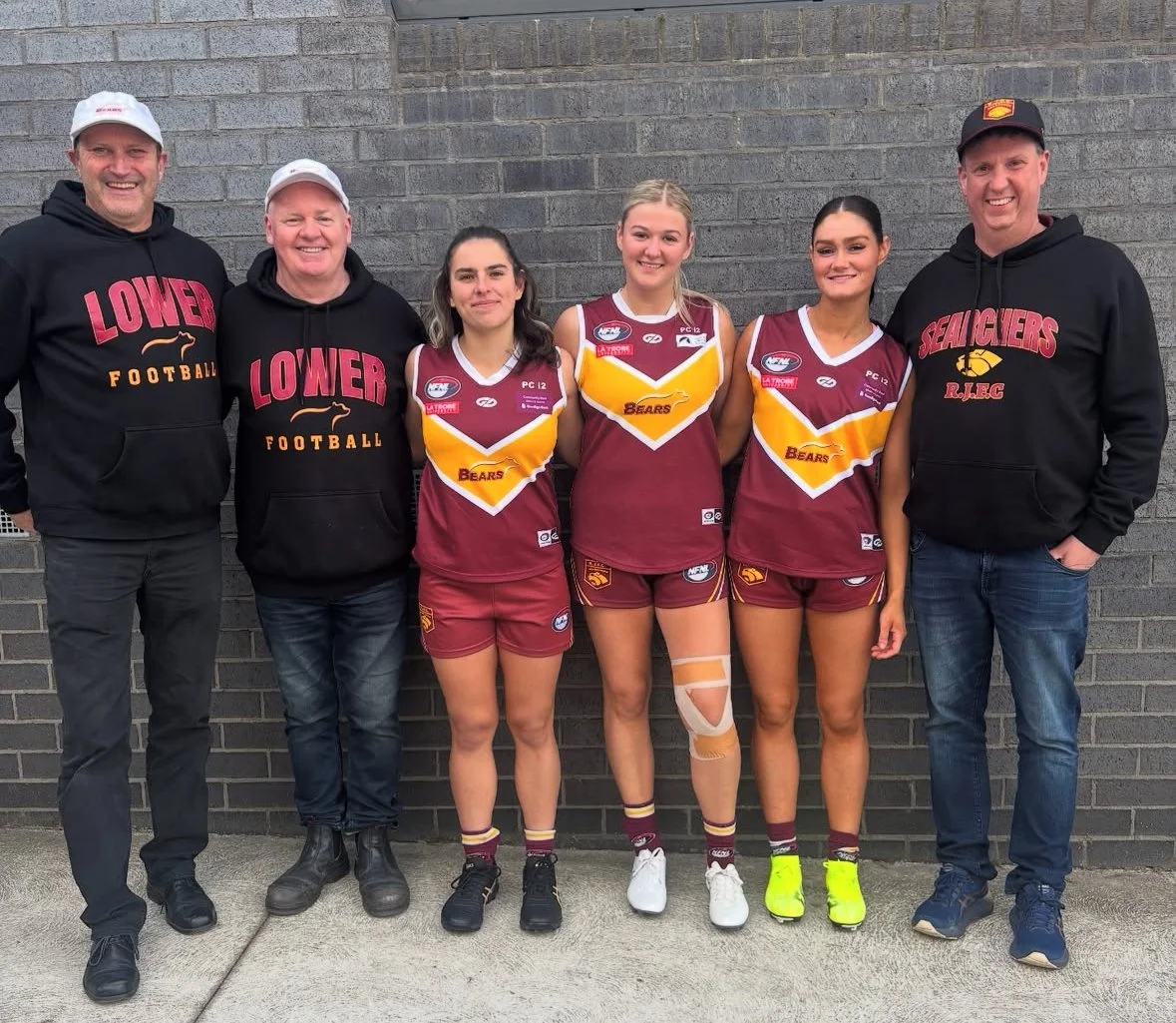 Group of six people, three men and three women, standing in front of a brick wall. The women are wearing maroon and yellow sports jerseys and shorts, likely a netball or basketball team. The men are dressed in casual attire with black hoodies and caps, with the words "Lower Football" and "Seagulls R.J.E.C" on the hoodies.