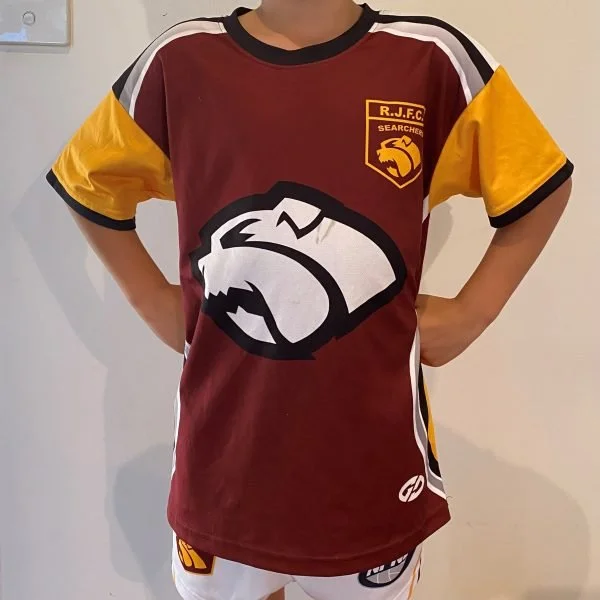 A person wearing a maroon sports jersey with yellow and white accents, featuring a large graphic of an bulldogs head and the logo of R.J.F.C. SEARCHERS on the chest.