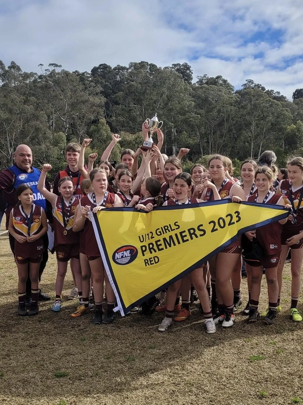 Girls' under 12 sports team celebrating victory with a trophy, holding a banner that reads 'U/12 Girls Premiers 2023, AFL', outdoors on a grassy field with trees in the background.