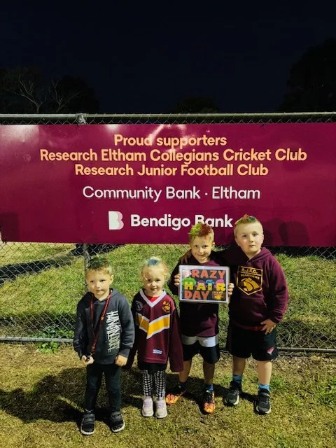 Four children standing in front of a purple banner promoting the Research Eltham Collegeians Cricket Club and Junior Football Club, with logos of Community Bank Eltham and Bendigo Bank at night.