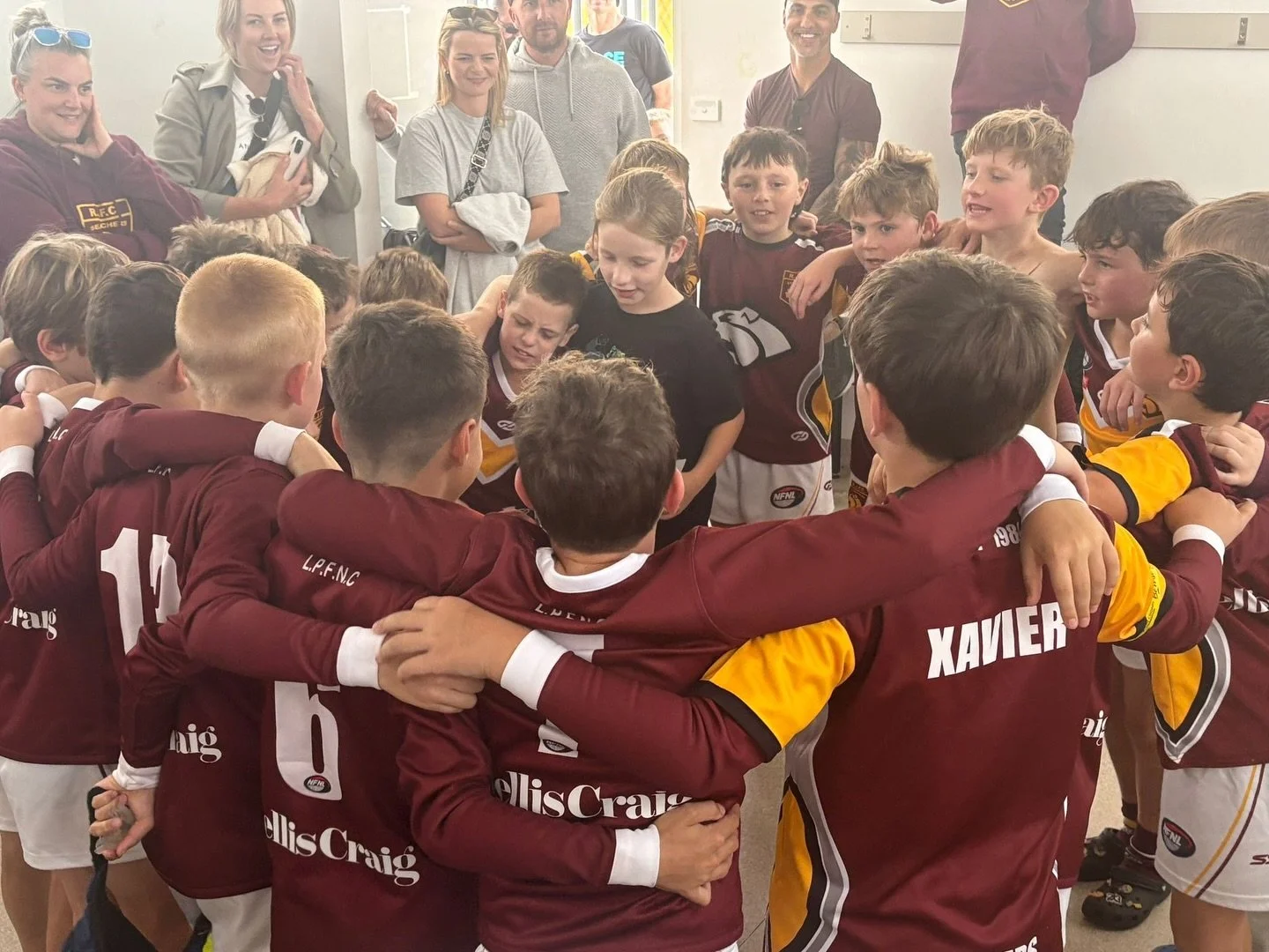 Young athletes in maroon jerseys huddle together in a circle with arms around each other, in a team pep talk, while a group of adults watch and smile in the background.