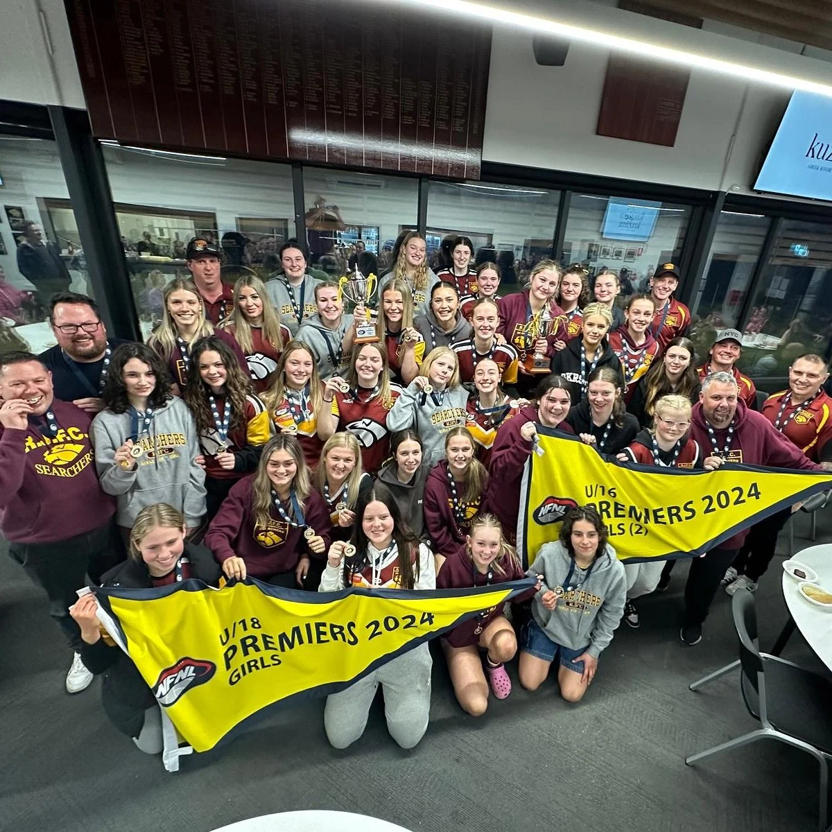 A large group of young female sports players and coaches posing together at an indoor sports venue, holding medals, trophies, and banners that read "U/16 PREMIERS 2024 GIRLS" and "U/18 PREMIERS 2024 GIRLS."