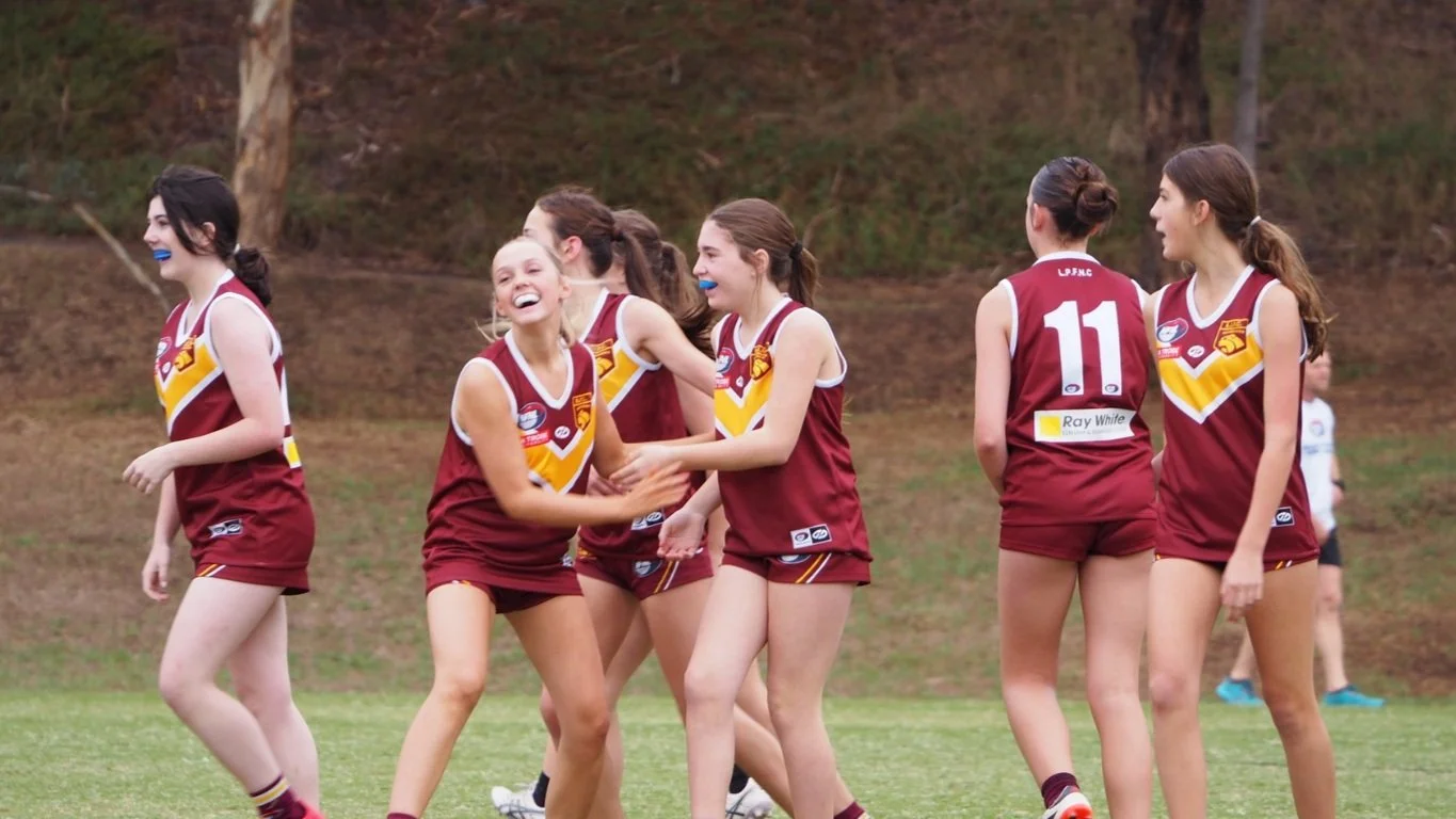A group of female athletes in maroon and yellow sports uniforms celebrating on a grassy field