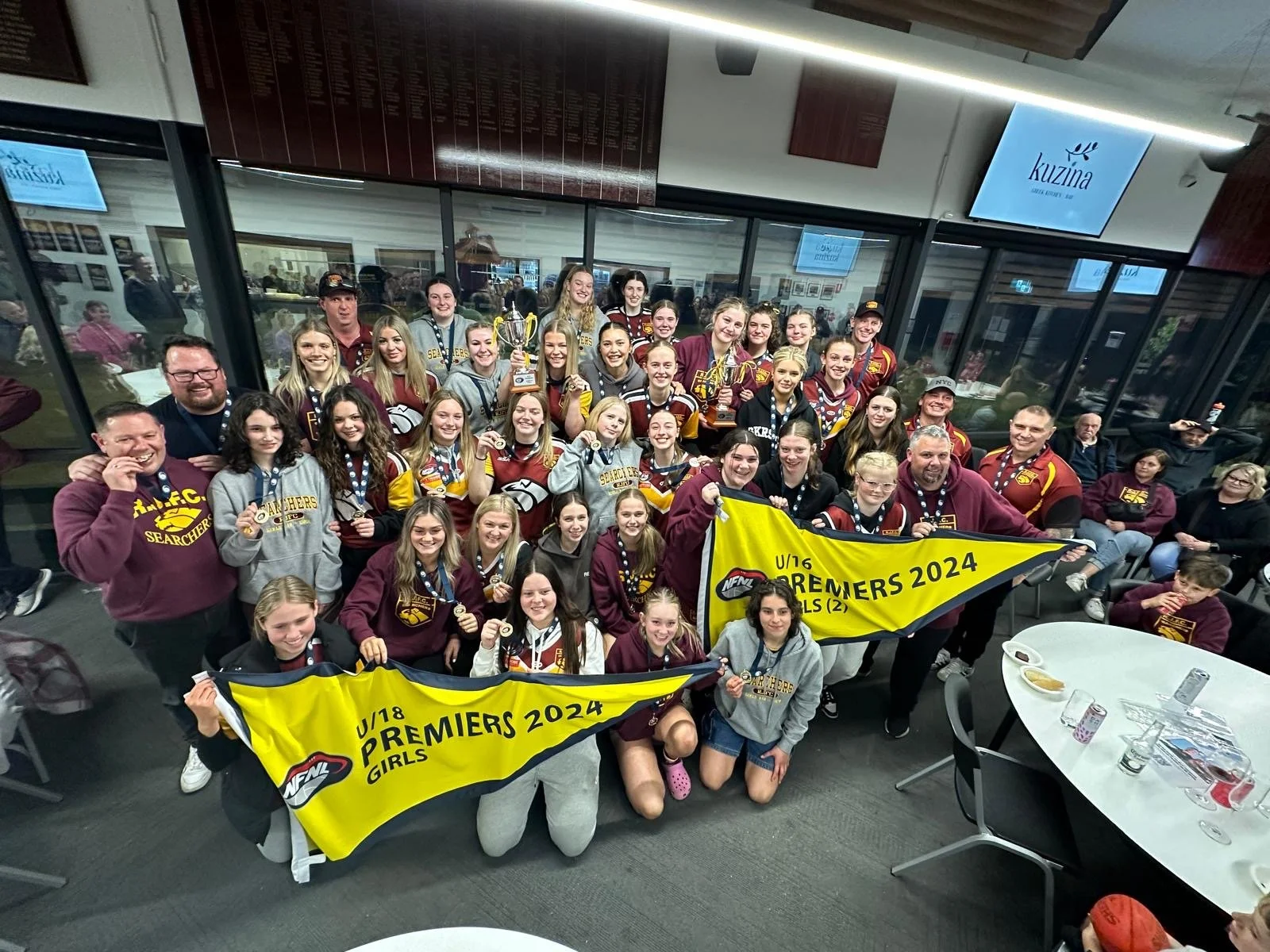 A large group of young female athletes and their coaches celebrating their victory at an indoor sports event, holding medals, trophies, and yellow banners that read "U/18 Premiers 2024 Girls" and "U/16 Premiers 2024 Girls."