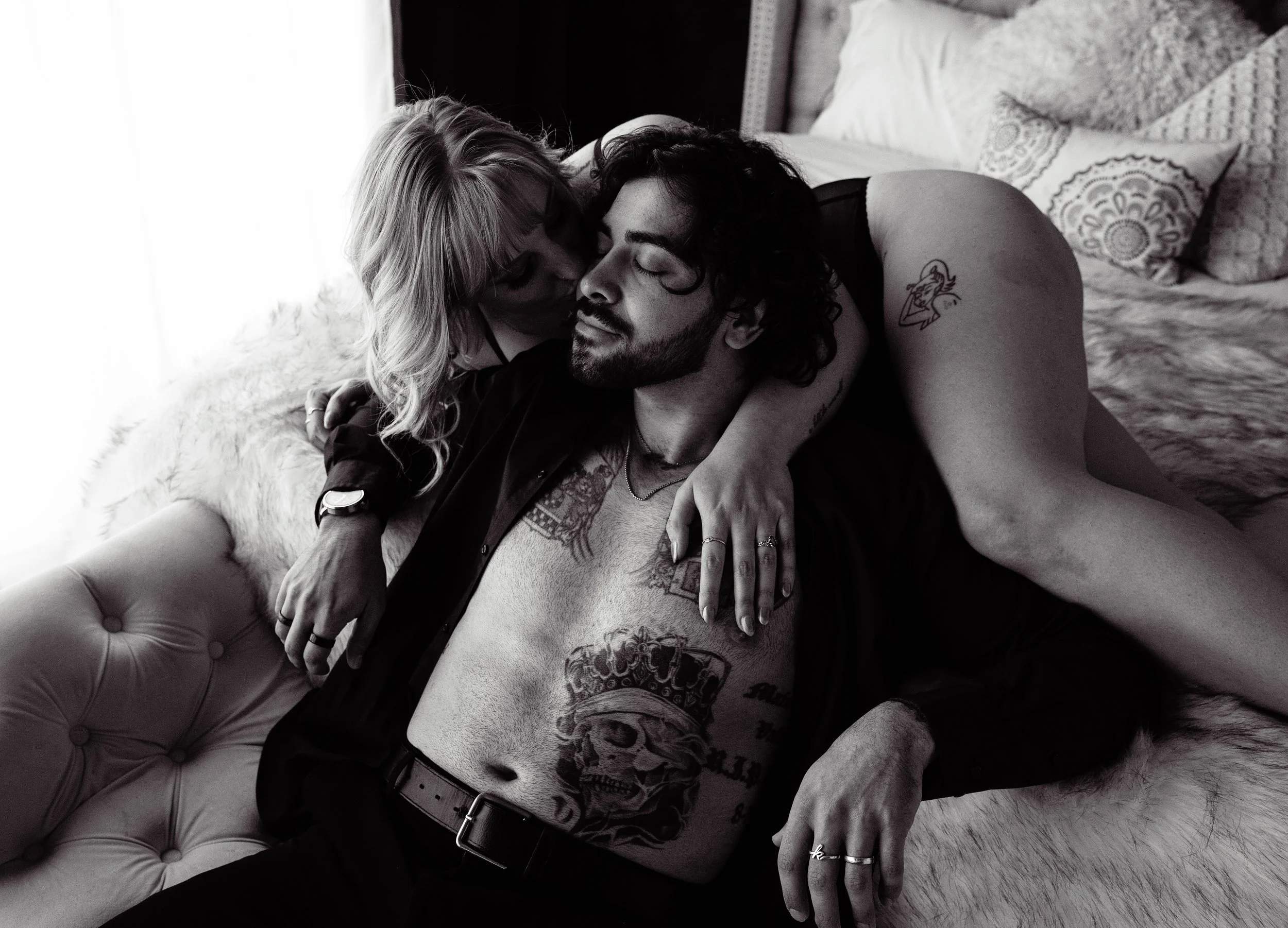 Boudoir Photo: A black and white photo of a couple lying on a bed, sharing an intimate moment. The woman is draped over the man, kissing his cheek, while the man has his eyes closed and a slight smile.