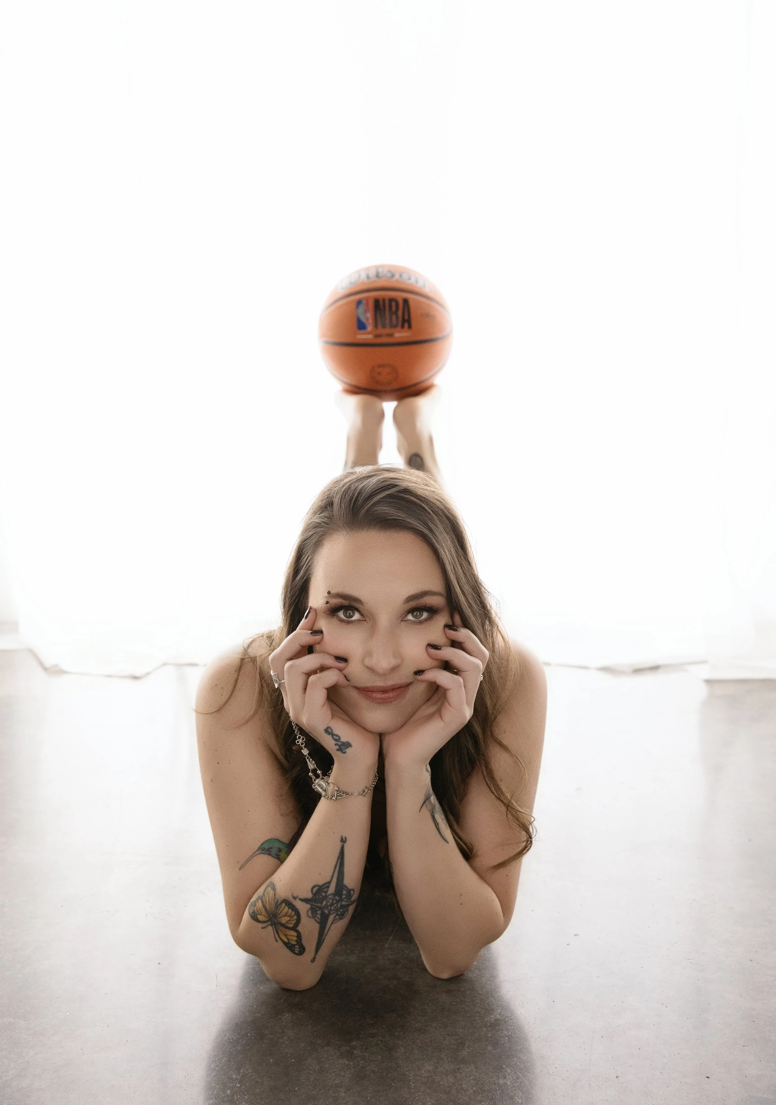 Boudoir Photo: Young woman with tattoos lying on her stomach on the floor, resting her chin on her hands, smiling at the camera, with a basketball balanced on her feet raised above her for a shot.