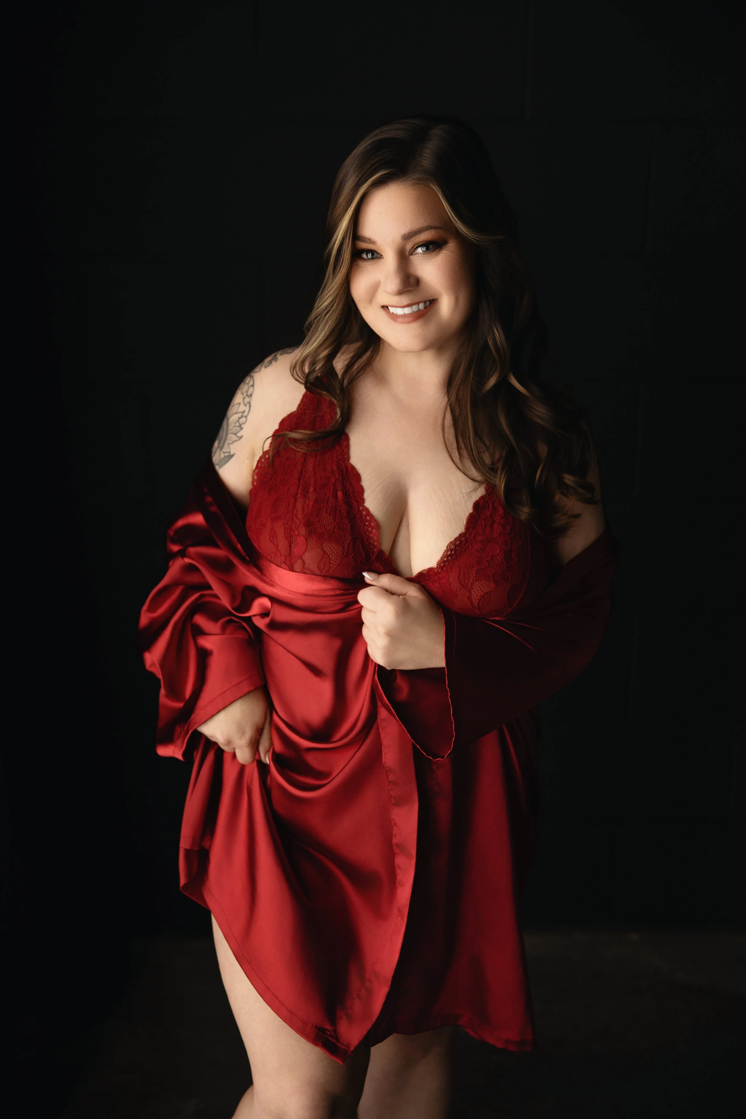 Boudoir Photo: Woman in red satin and lace lingerie with a silk robe, smiling and holding her robe open against a dark background.
