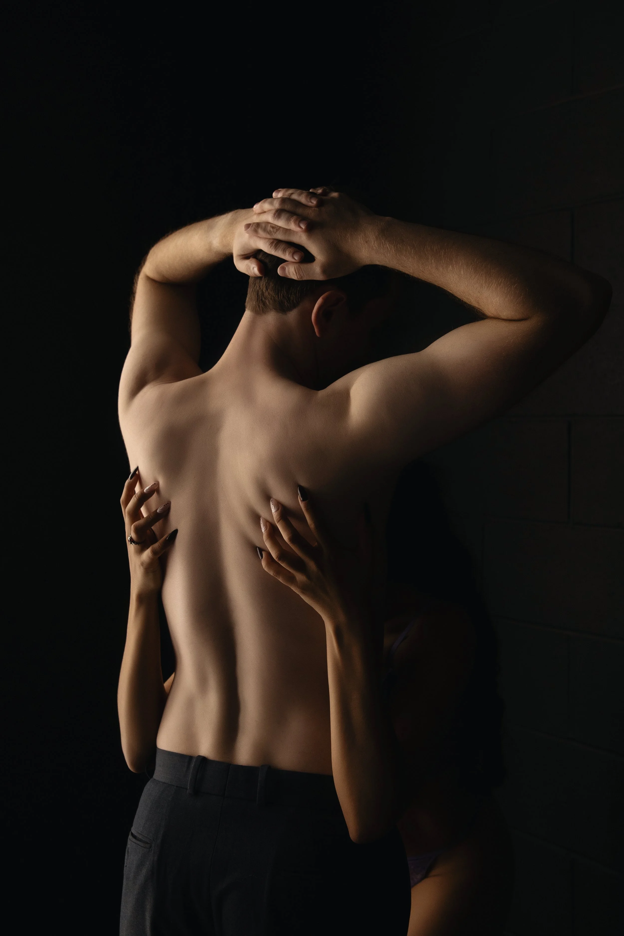 A shirtless person with short hair, with hands on their head, is being embraced from behind by another person with long nails, who is pressing their hands against the first person's back. They are standing against a dark background.