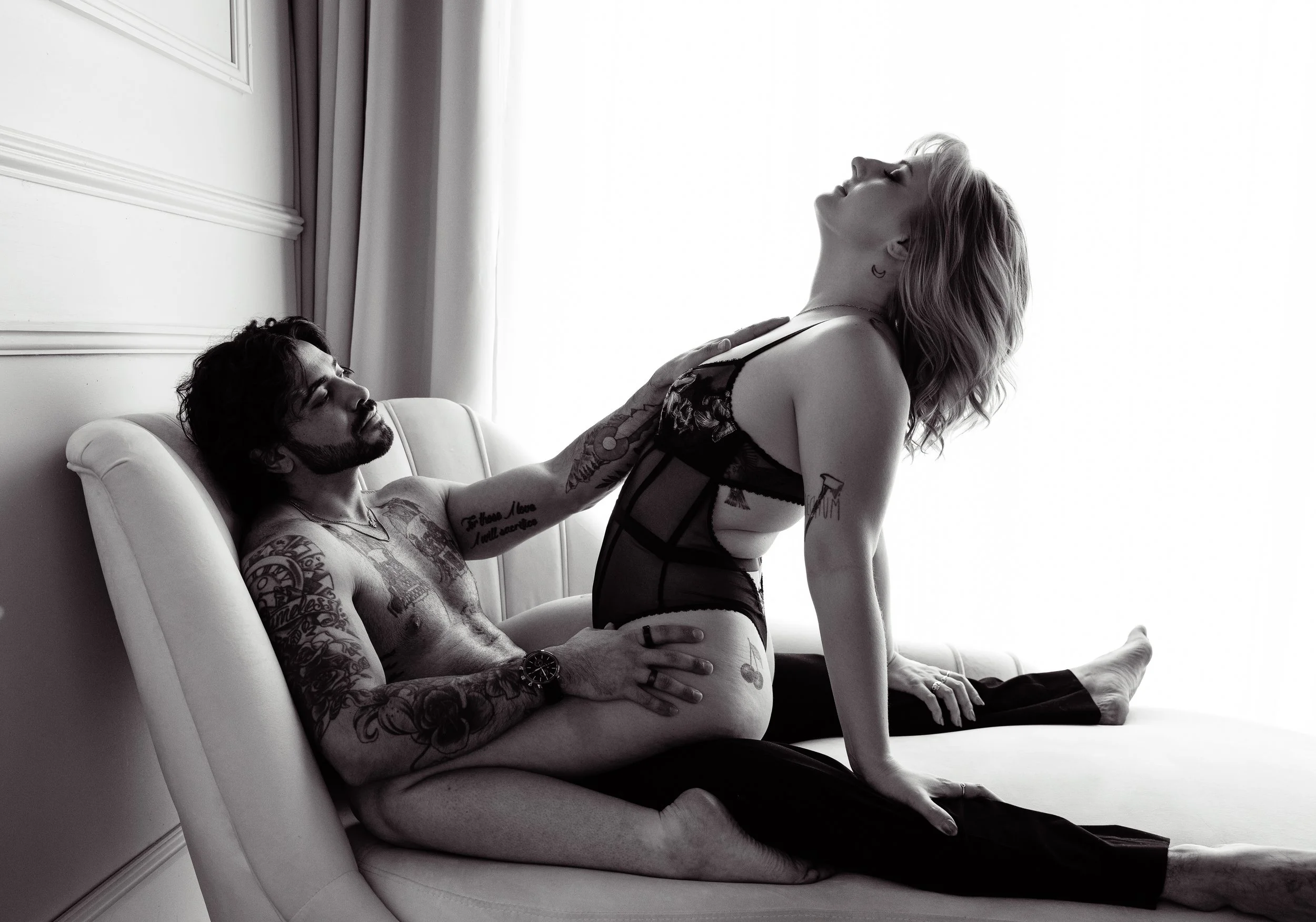 Boudoir Photo: A man and a woman are on a bed, with the man lying back against the headboard and the woman sitting on his lap. The woman, wearing lingerie, is leaning back with her head tilted upward, and the man is holding her waist.