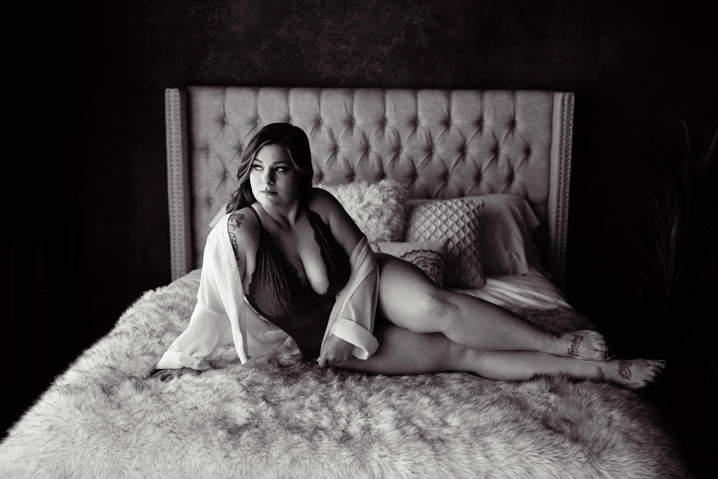 Boudoir Photo: A woman reclining on a bed with a fluffy blanket, wearing dark lingerie and a light open shirt, looking to her left in a room with a tufted headboard and pillows, in black and white.