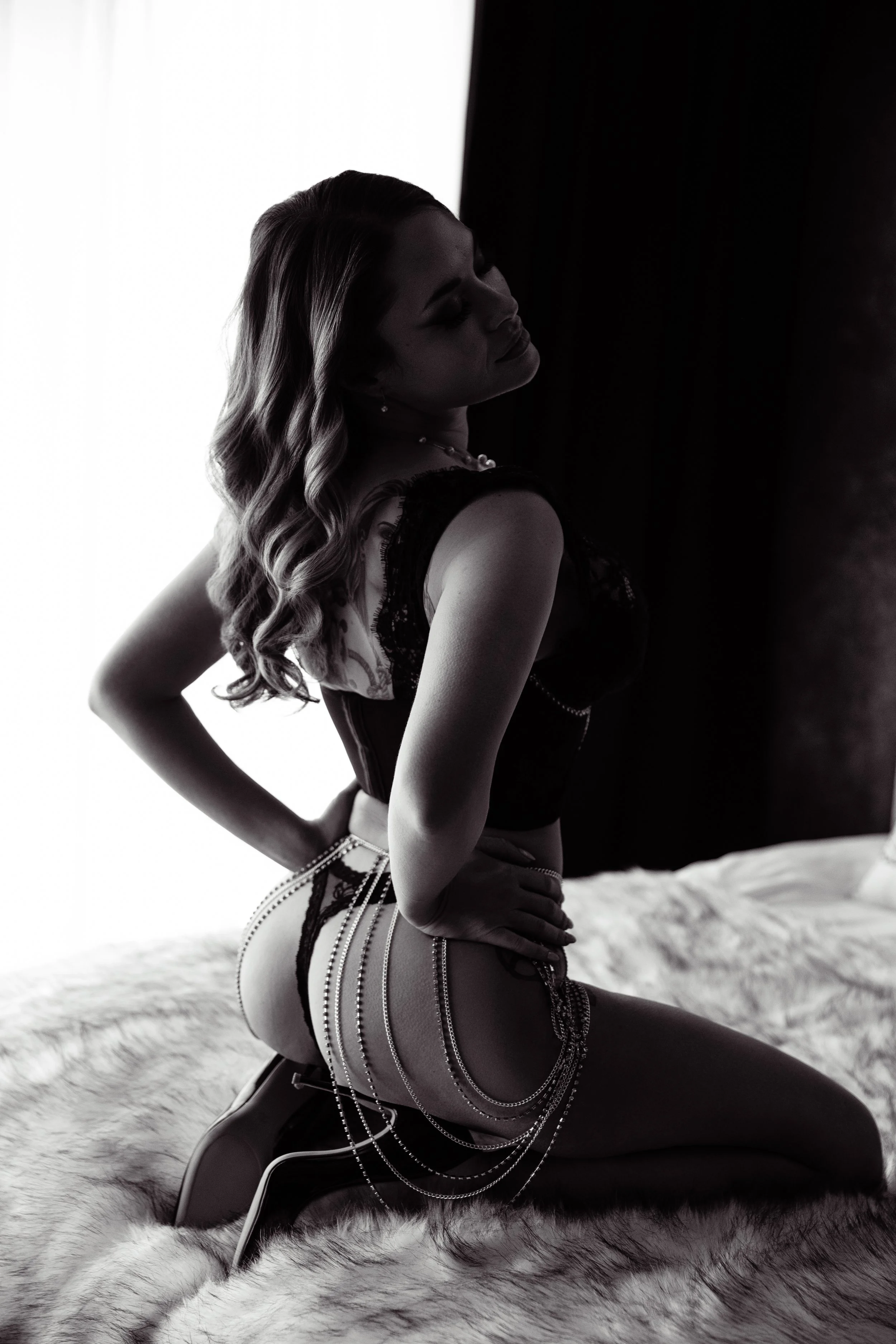 Boudoir Photo: A woman with styled hair kneels on a bed, wearing lingerie and jewelry, with her eyes closed and head tilted back, in a dimly lit room.