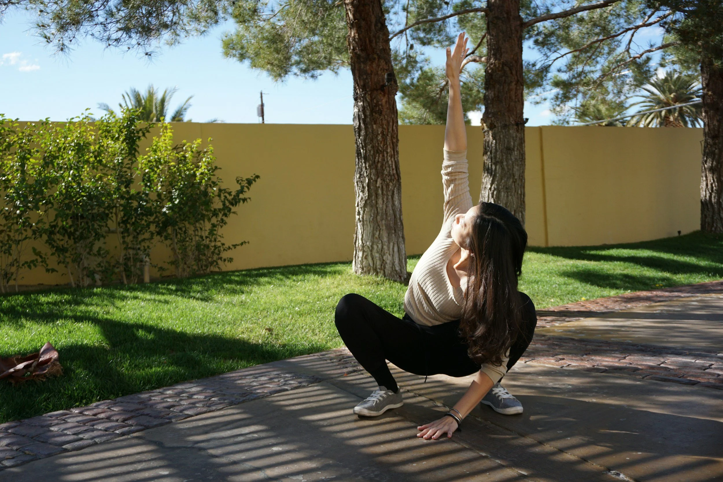 a woman in a deep squat with one hand reaching towards the sky outdoors on her back patio in fitness clothes and the morning sun