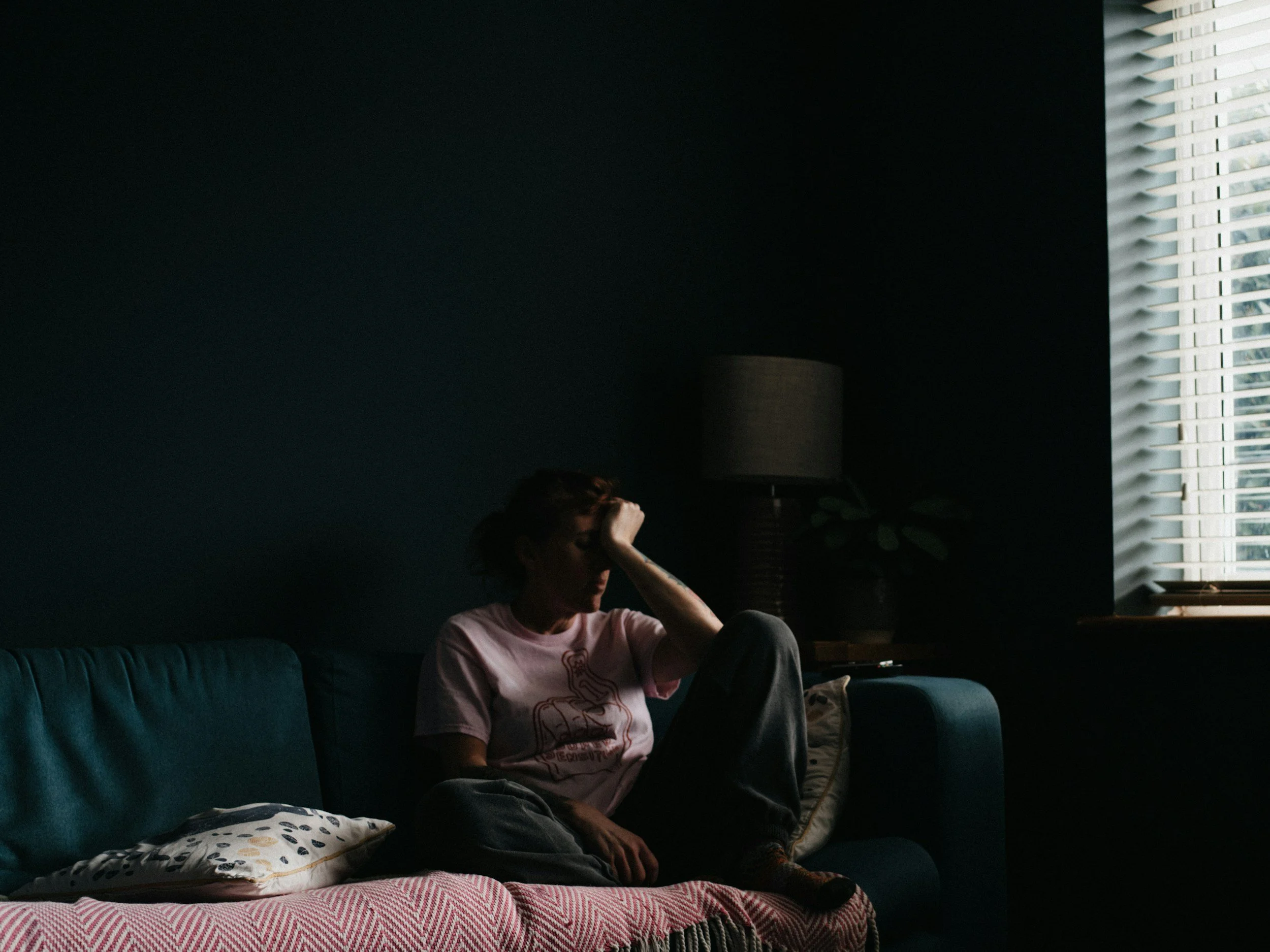 a tired woman with her head in a hand sitting on a couch with her legs up on the couch. she's wearing a big tee and sweats and the room is dark with light coming in through a single window and blinds