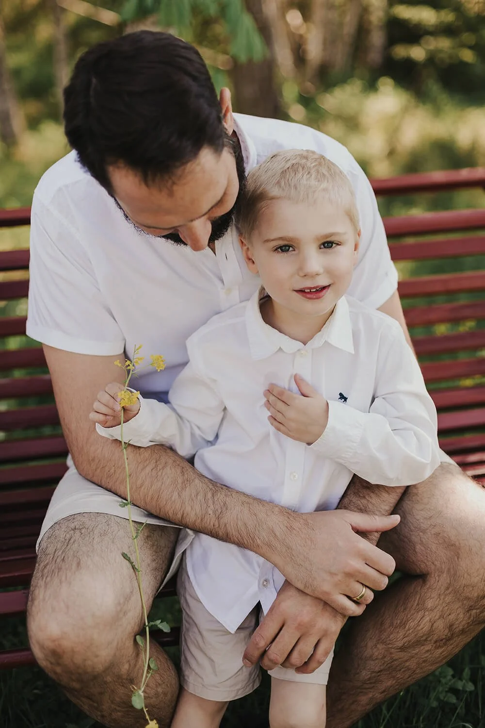 Et nært portrett av en far og sønn som sitter sammen på en rød trebenk utendørs på Konnerud. Faren ser ned på gutten, som holder en liten gul blomst og ser mot kameraet. Begge er kledd i hvite skjorter i naturlig dagslys.