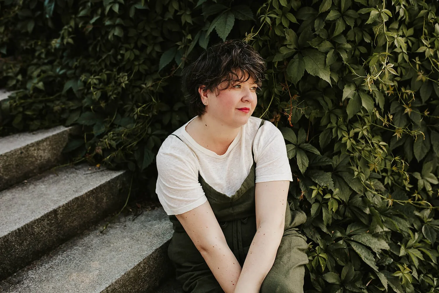En kvinne med mørkt hår som sitter i en steintrapp foran en tett vegg av grønne blader i Fajanseparken, Drammen.