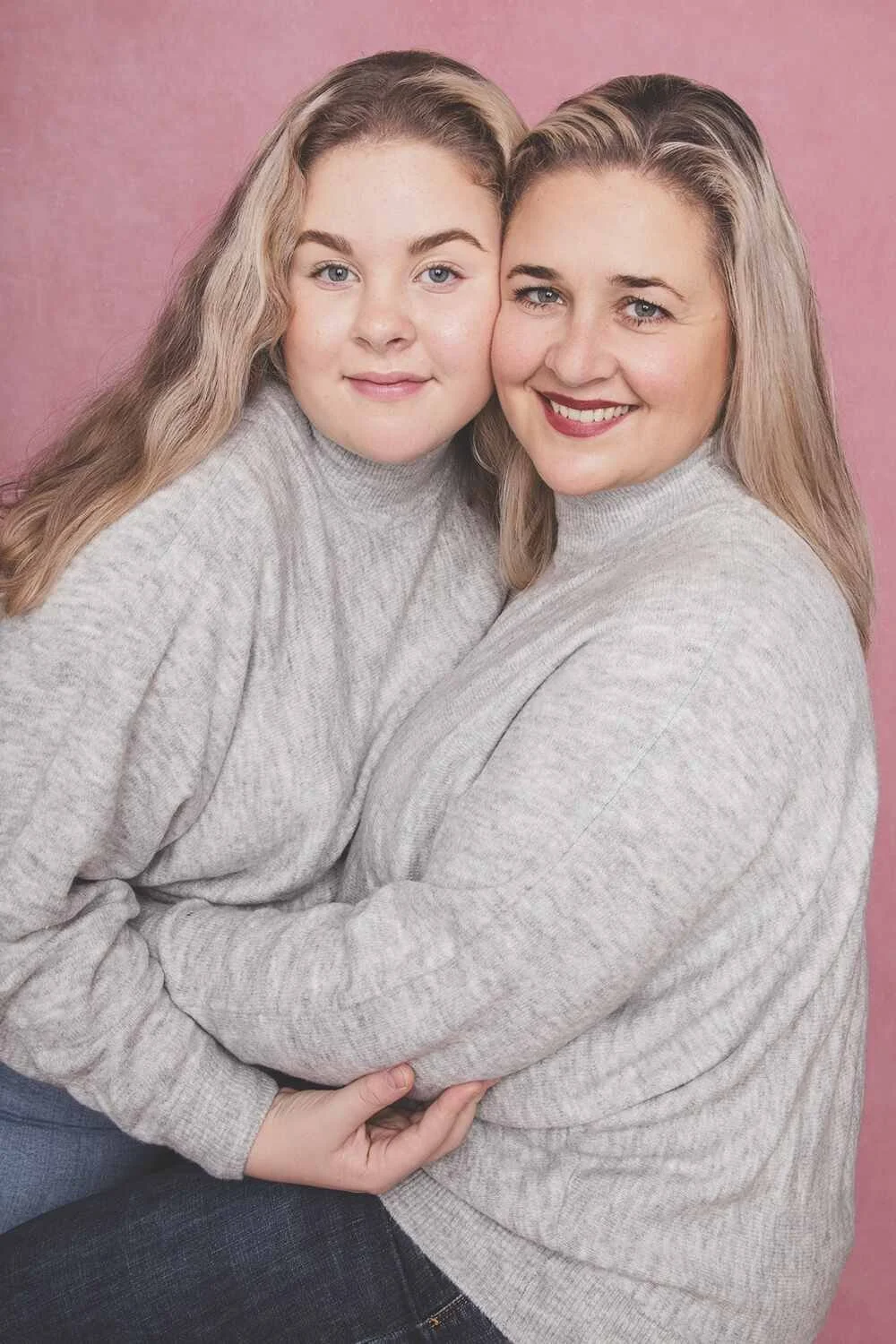 To kvinner med lyst hår, smilende, kledd i lyse grå gensere, står tett sammen mot en rosa bakgrunn, og smiler mot kamera.