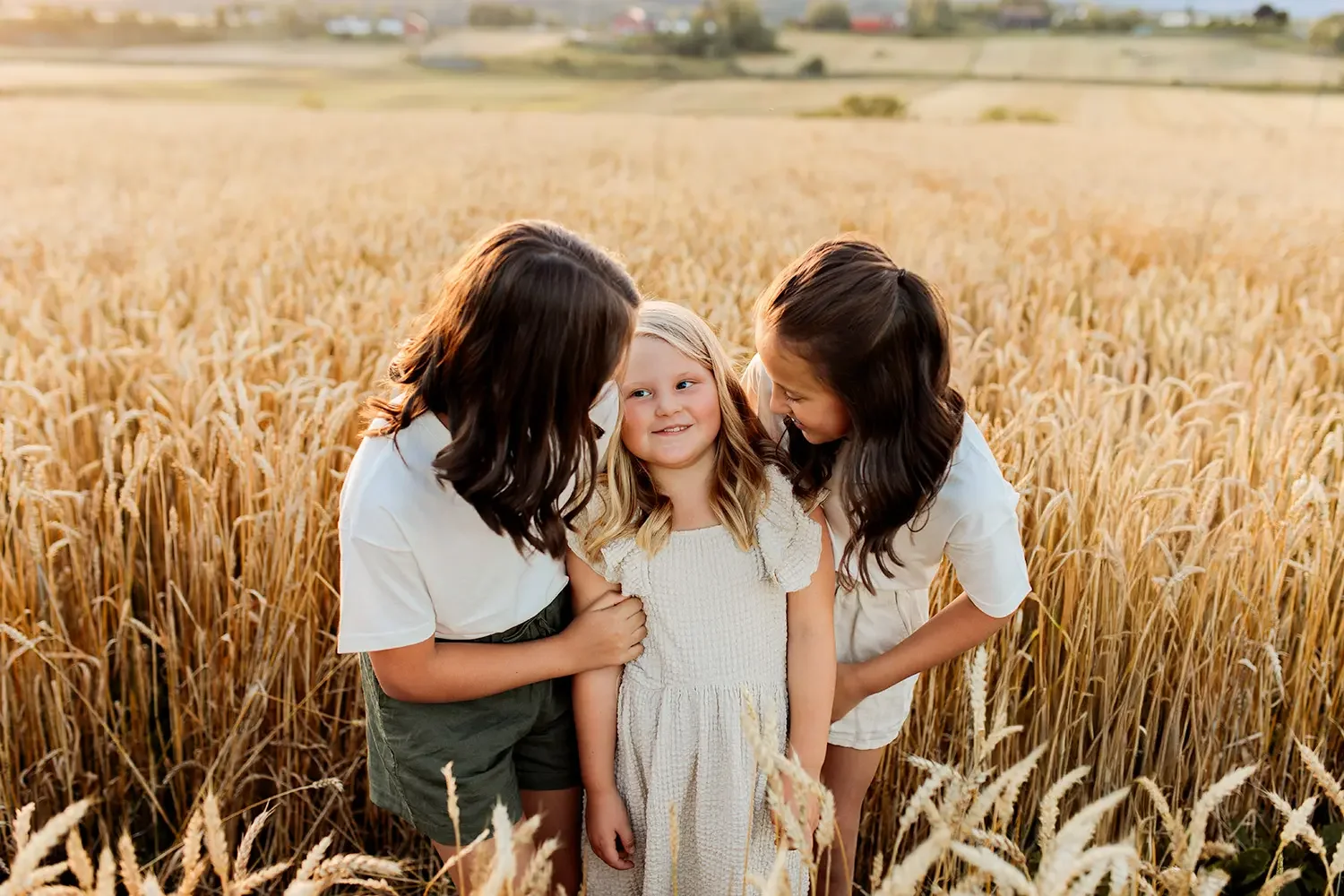 Tre søstre står tett sammen i en gyllen åker under en familiefotografering under golden hour.