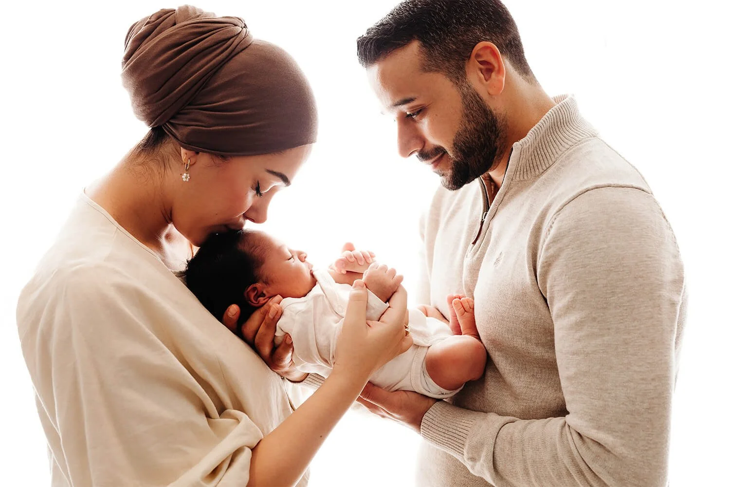 Et lyst og hjertevarmt bilde fra en nyfødtfotografering der to foreldre holder sin nyfødte baby sammen. Mor kysser babyen ømt på pannen i et lyst rom med naturlig dagslys. Bildet er tatt hjemme i Solbergelva.