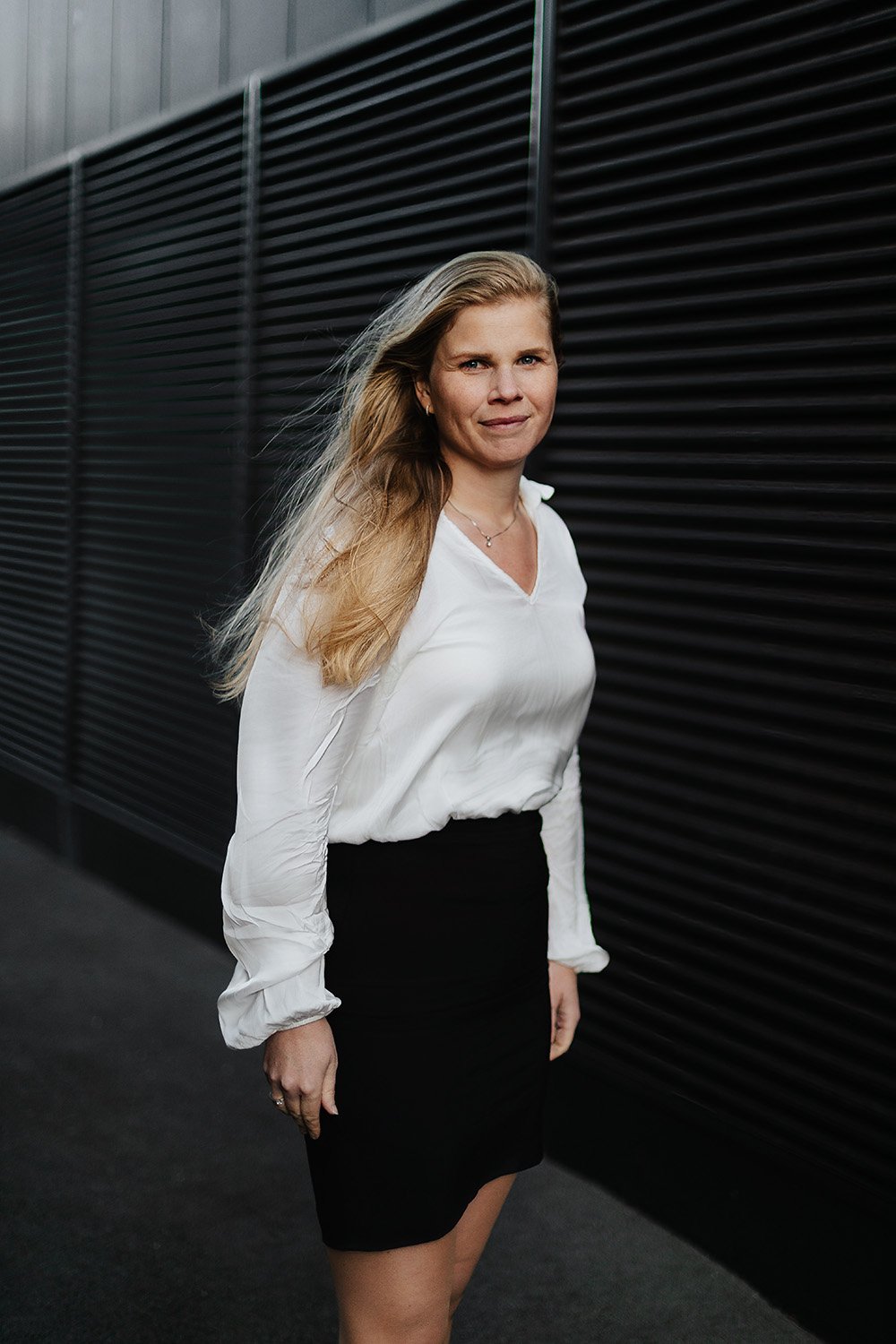 Portrett av en kvinne i hvit overdel som står foran en moderne, mørk fasade på Union Brygge.