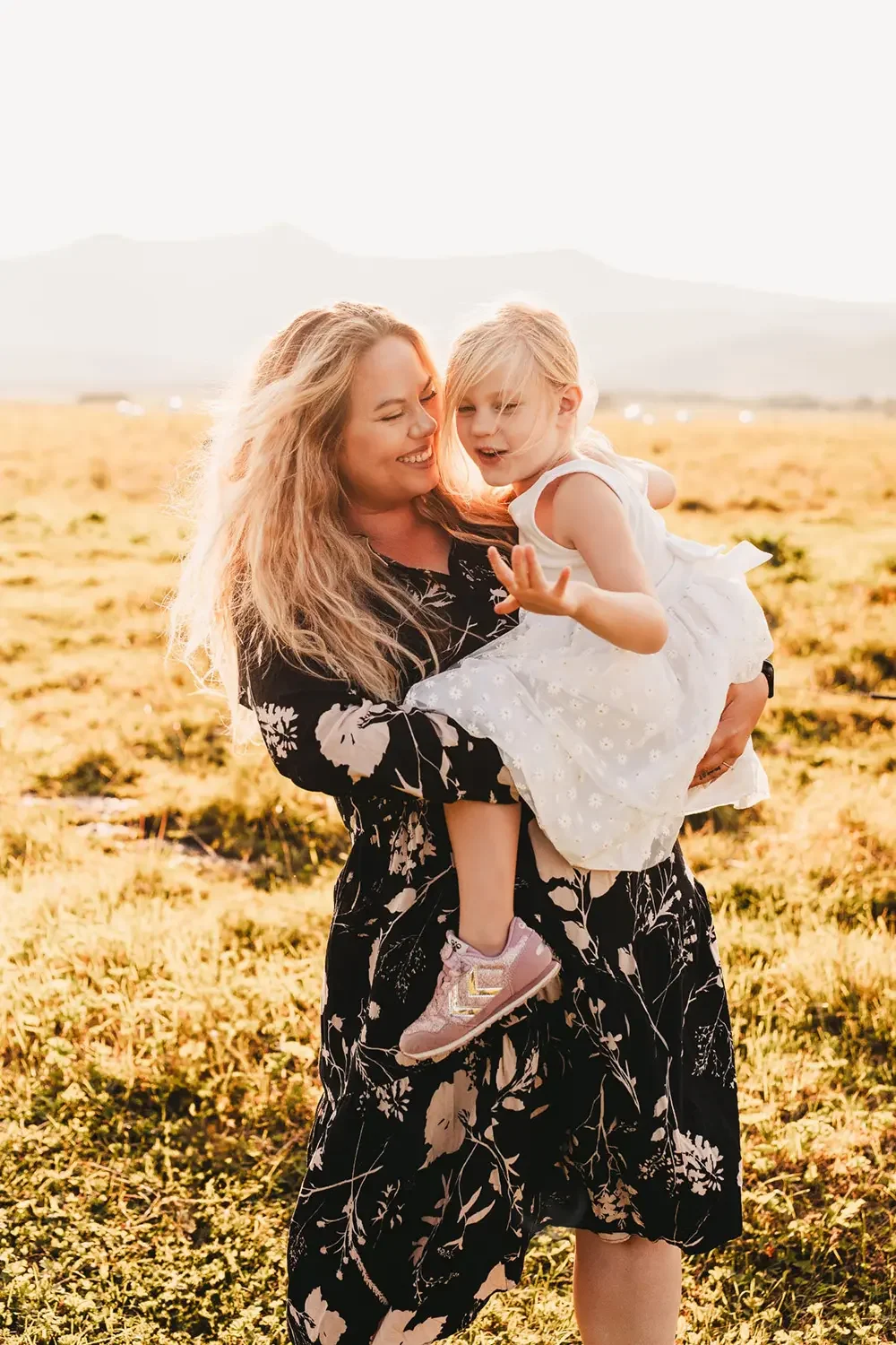 En smilende mamma som holder datteren sin på en åpen slette på Andøya under solnedgangen. I bakgrunnen ser man majestetiske fjell i det gylne lyset. Bildet er tatt av fotograf Tina Bergersen fra Drammen.