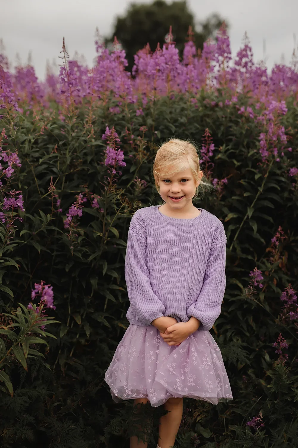 Jente som smiler med lilla genser og skjørt foran lilla blomster