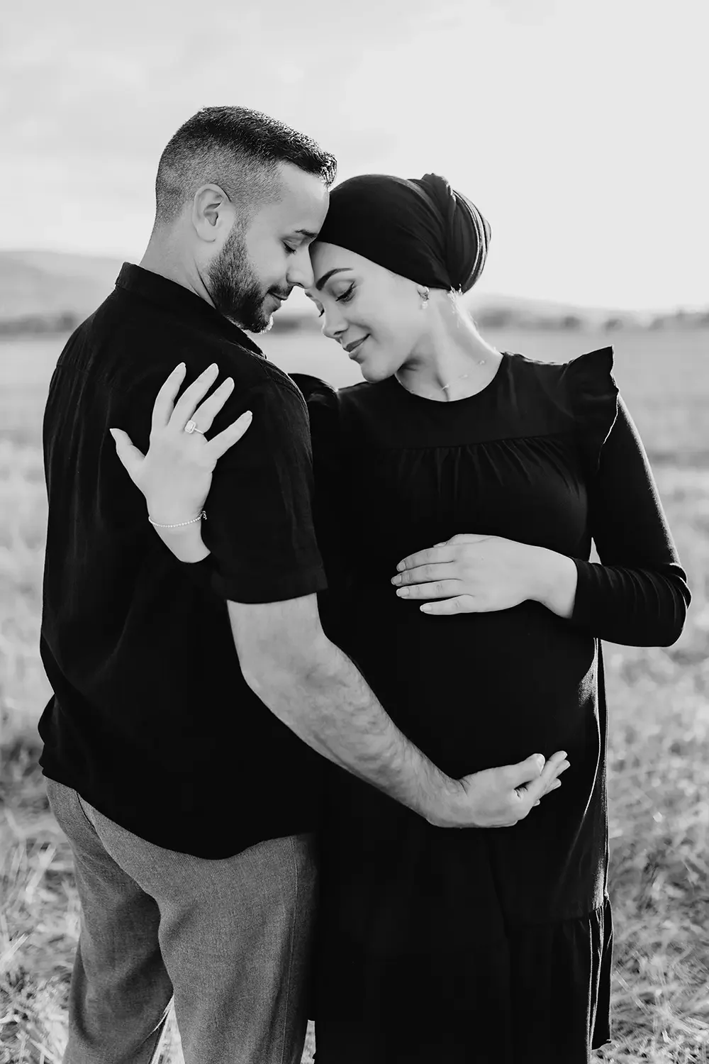Et vakkert svart-hvitt bilde av et par som står tett sammen på en åker i Solbergelva, Drammen. Kvinnen er gravid og bærer hodeplagg, og begge holder hendene kjærlig på den gravide magen mens de lener pannene mot hverandre.