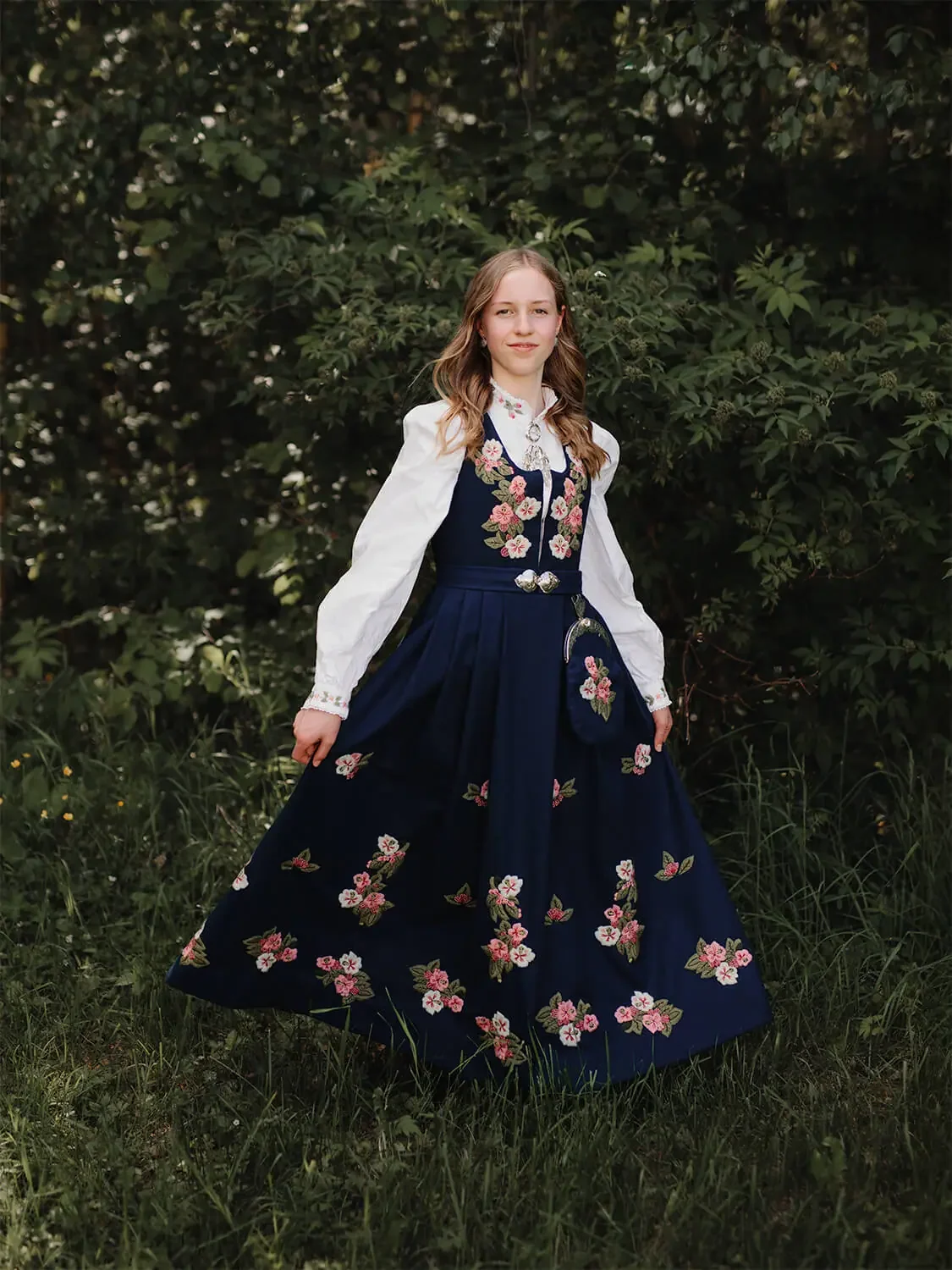 En stående konfirmant i en blå Lier-bunad utendørs i grønne omgivelser på Konnerud i Drammen. Hun holder skjørtet litt ut til siden og smiler til kameraet.