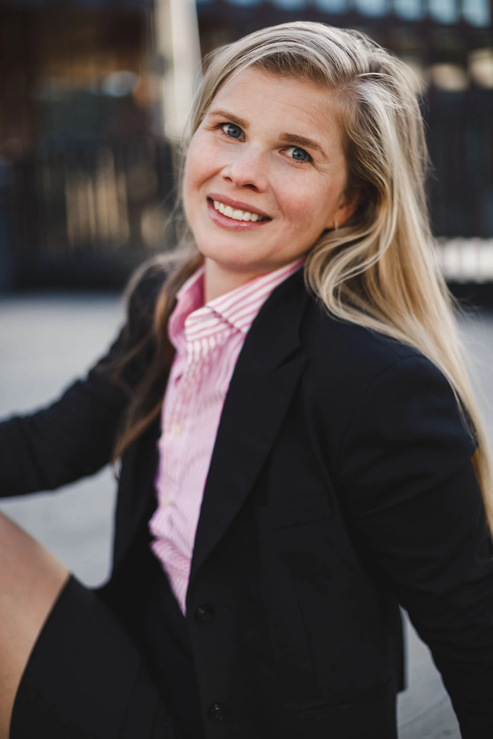En kvinne med blondt hår, iført svart blazer og rosa stripete skjorte, smiler og ser mot kamera i utendørs setting.